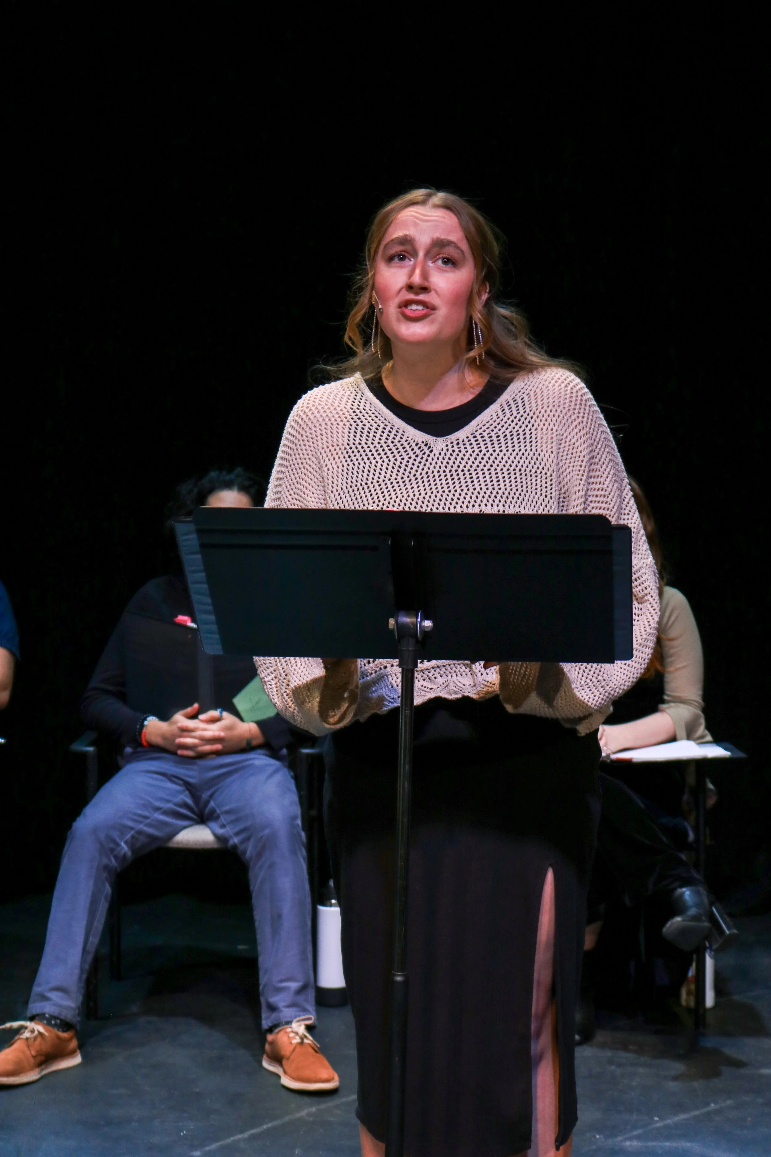 A woman is standing behind a music stand on a stage, speaking or performing, with several seated individuals in the background.