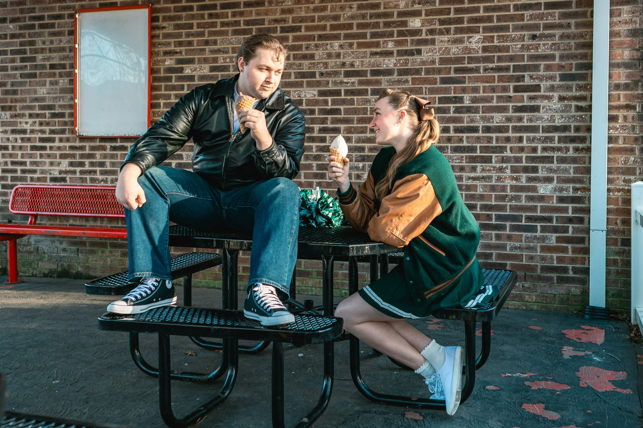 A young man and woman sitting outside on a black metal picnic table, sharing ice cream cones, with a brick wall in the background.
