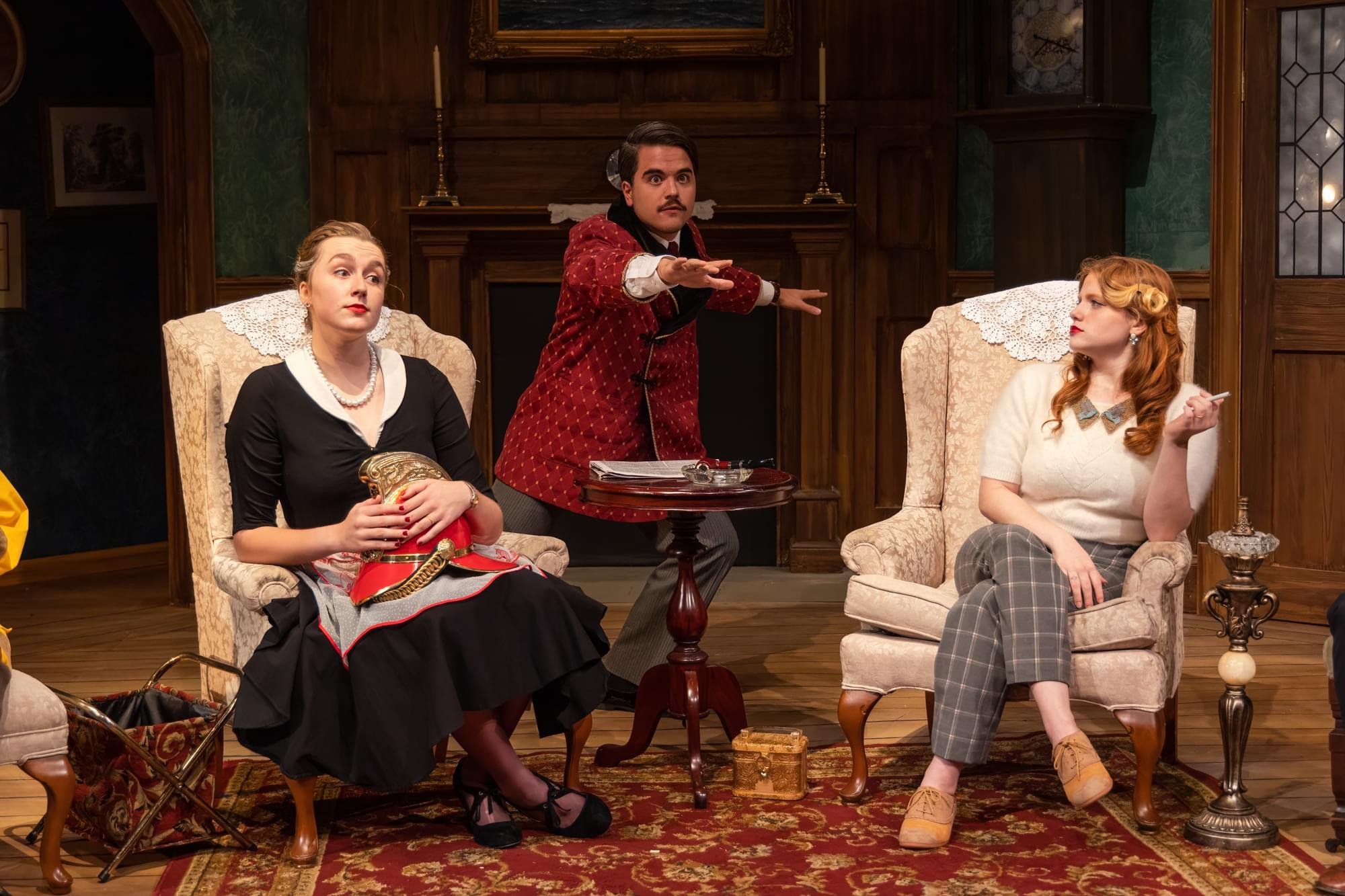 Three actors on stage, two women seated in armchairs and a man standing between them, in a vintage-style room. The woman on the left has blonde hair, wearing a black dress with a white collar, holding a sword. The man in the center has dark hair and a mustache, wearing a red patterned jacket. The woman on the right has red hair, wearing plaid trousers and a white sweater, holding a cigarette.