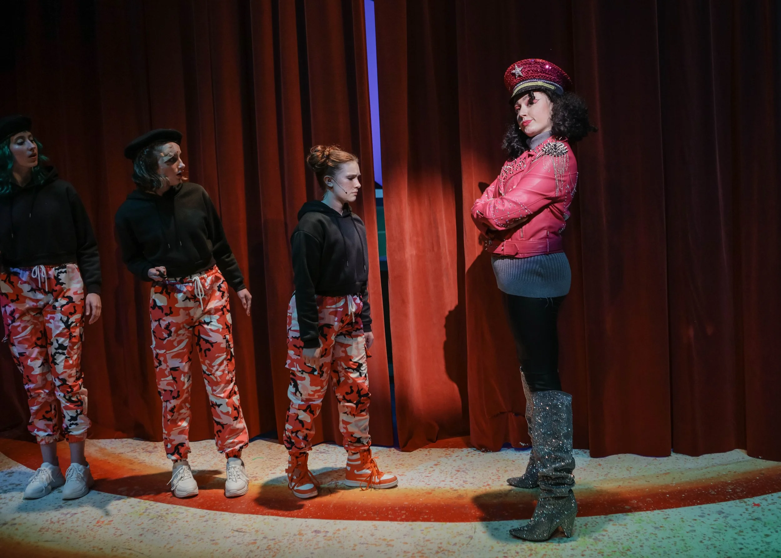 Four performers on stage, three young women wearing black hoodies and red camouflage pants, looking at a woman dressed in a pink jacket with silver studs, a sparkly red hat, and glittery silver boots, against a wooden curtain background.