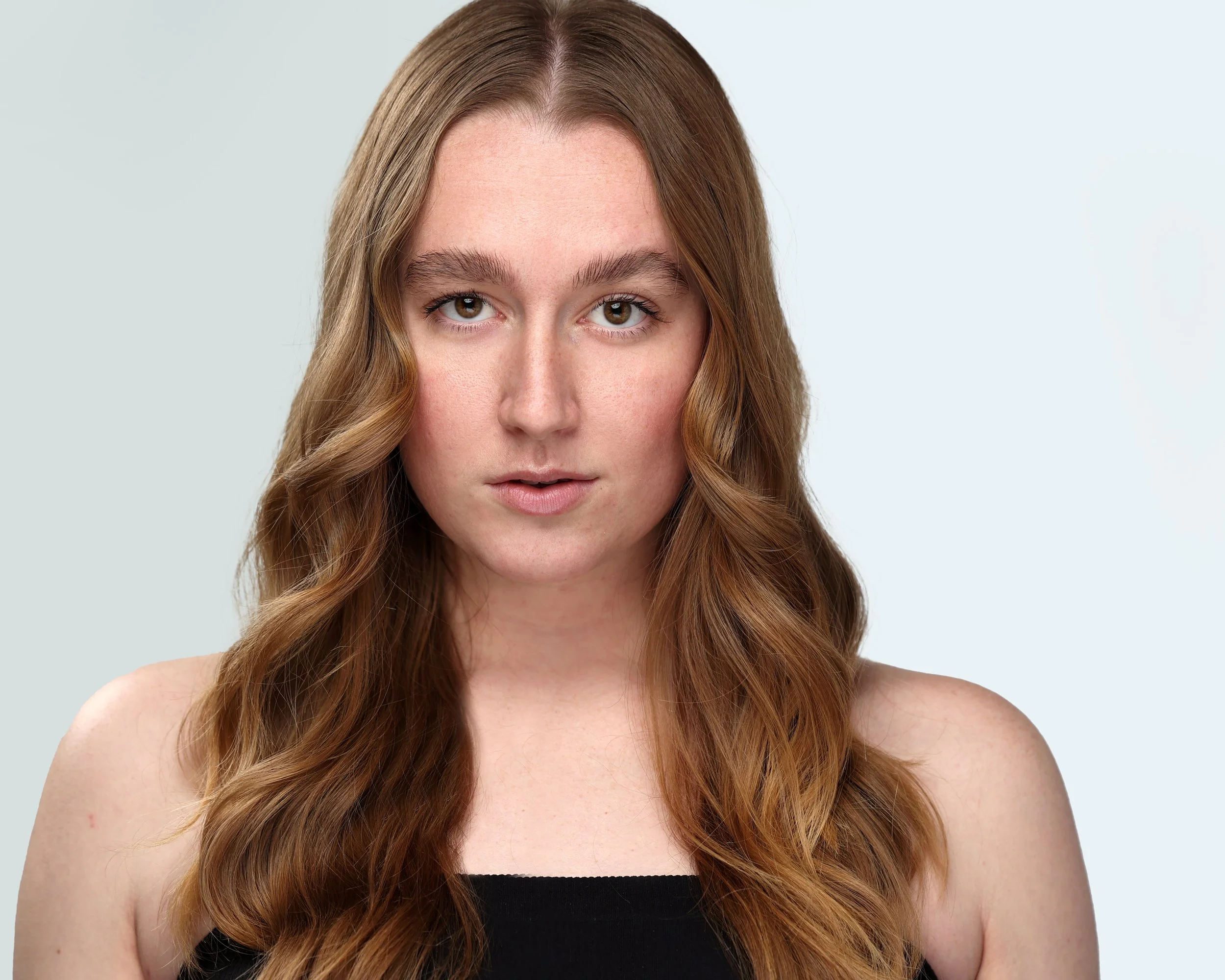 A young woman with long wavy red hair and fair skin, wearing a black top, looking directly at the camera with a neutral expression.