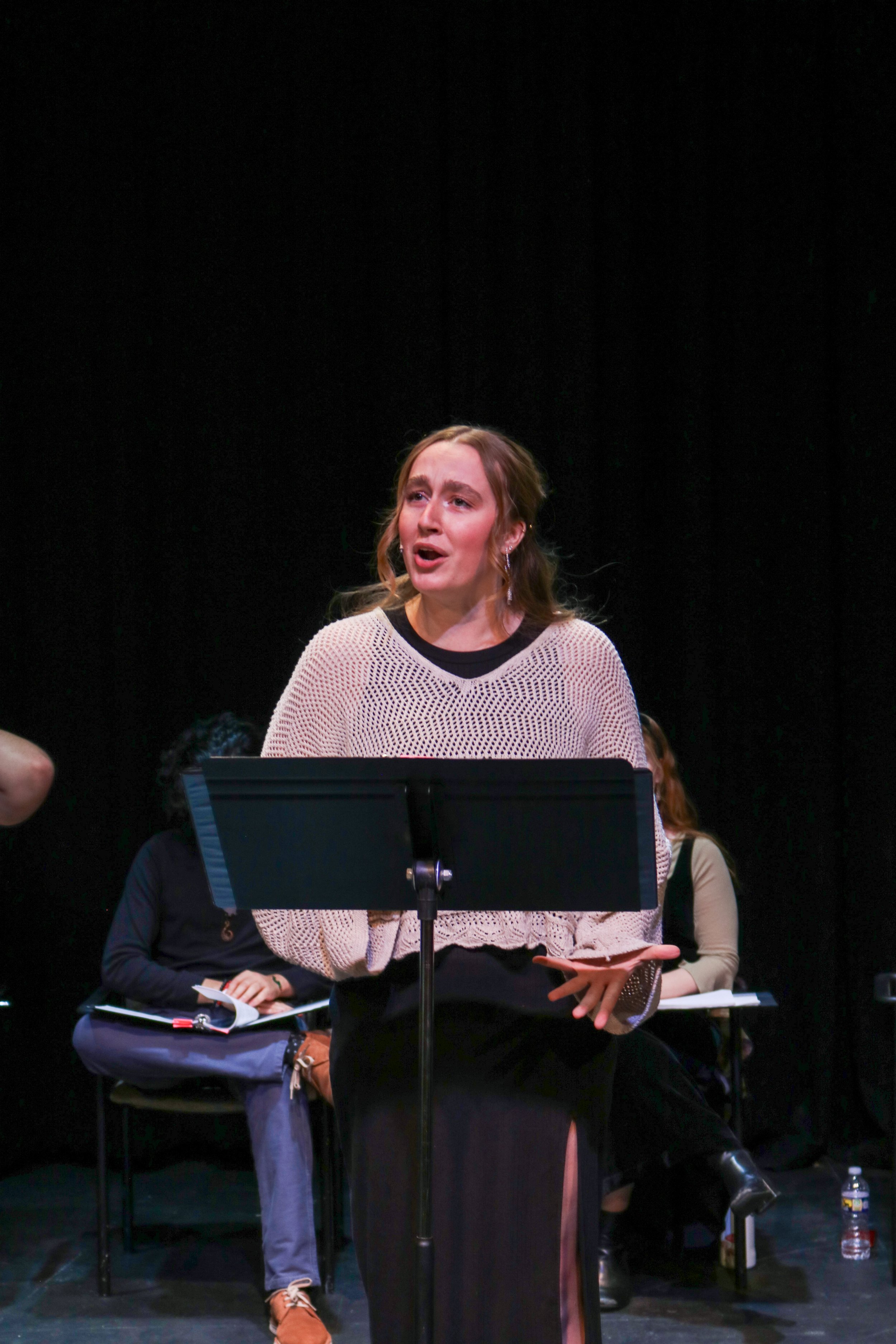 A young woman with light brown hair singing at a music stand during a performance, with others seated behind her on a dark stage.