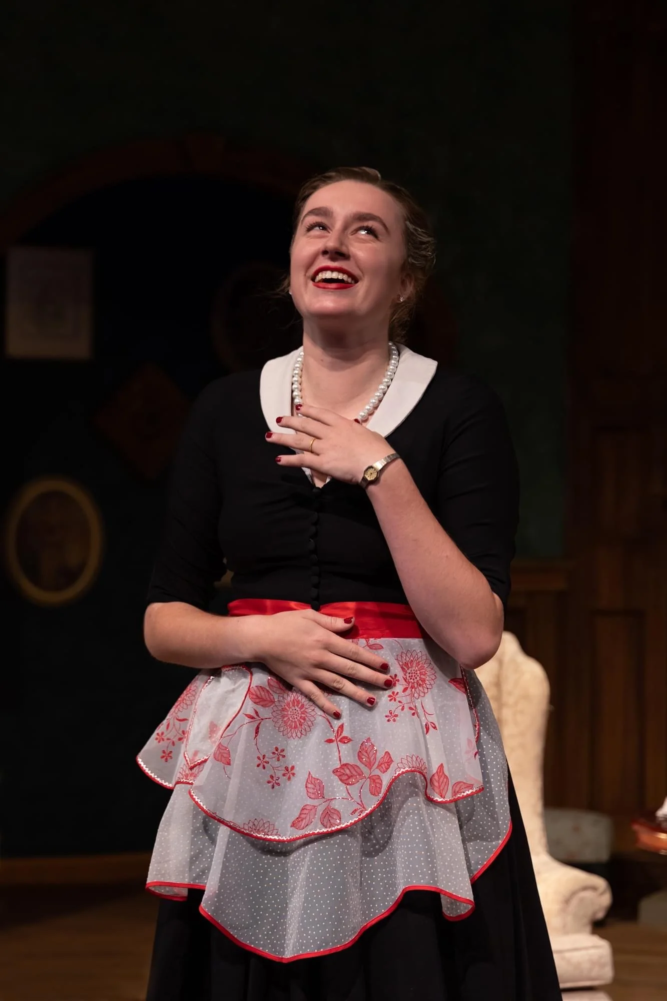 A woman with red lipstick and pearl necklace, dressed in vintage clothing, smiling on stage with her hand on her chest and stomach.