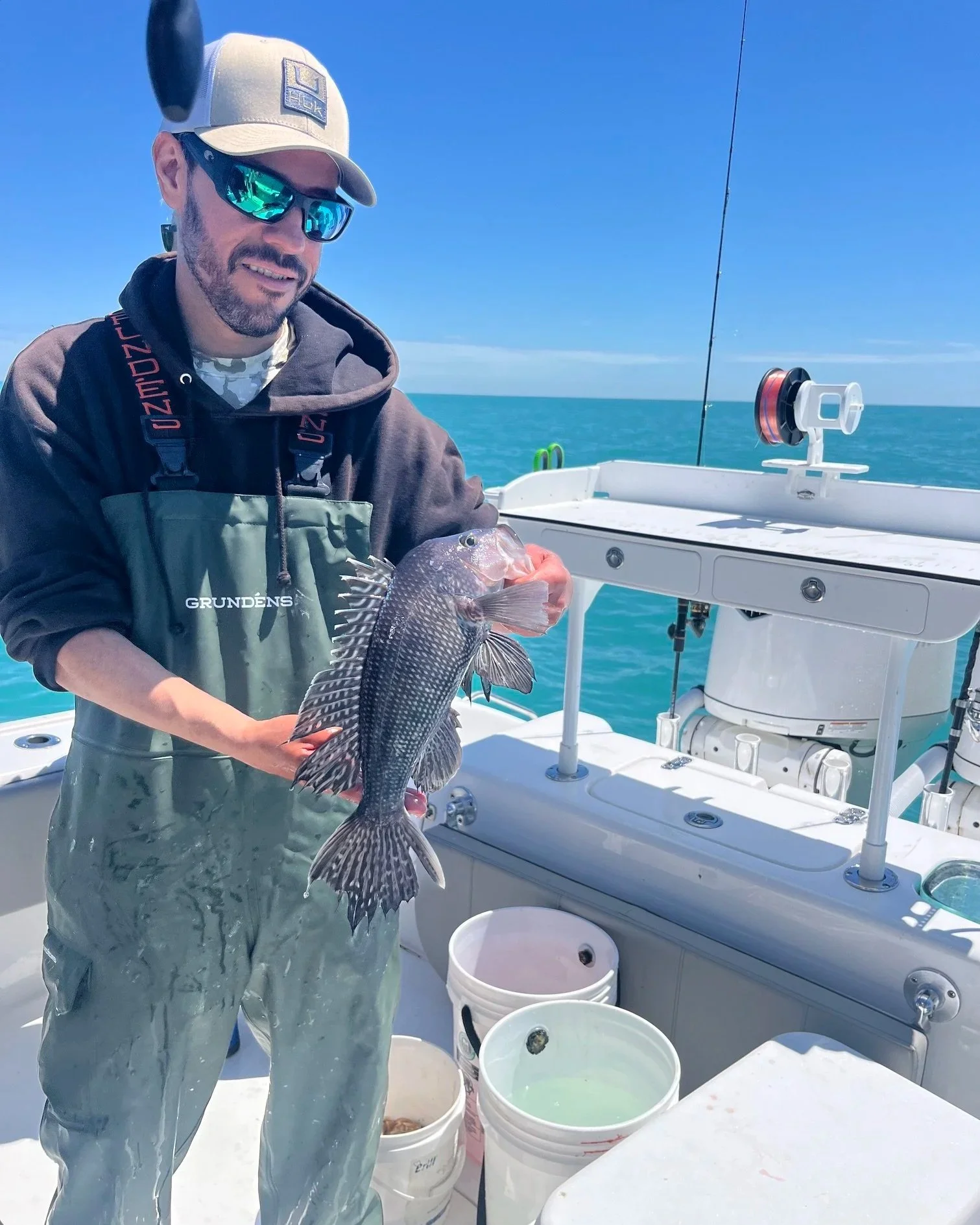 A man holding a striped fish on a boat in the open ocean under a clear blue sky, with buckets and fishing gear around him.