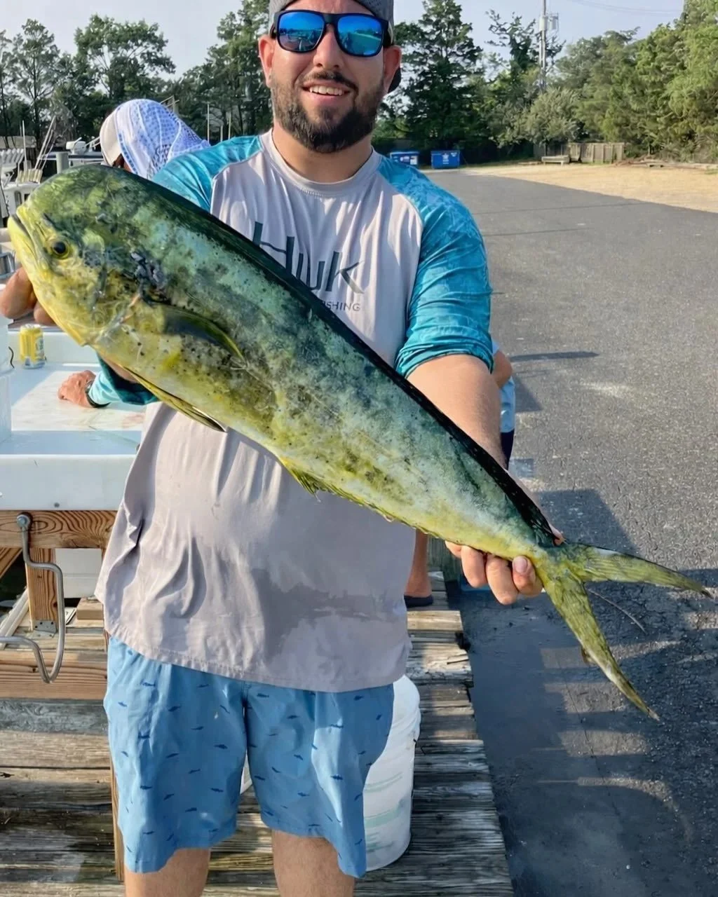 A man wearing sunglasses, a gray and blue rash guard, and blue swim trunks holding a large fish, likely a mahi-mahi, with a smile on his face on a dock or pier outdoors.