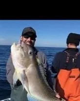 A man smiling and holding a large fish on a boat, with the ocean in the background.