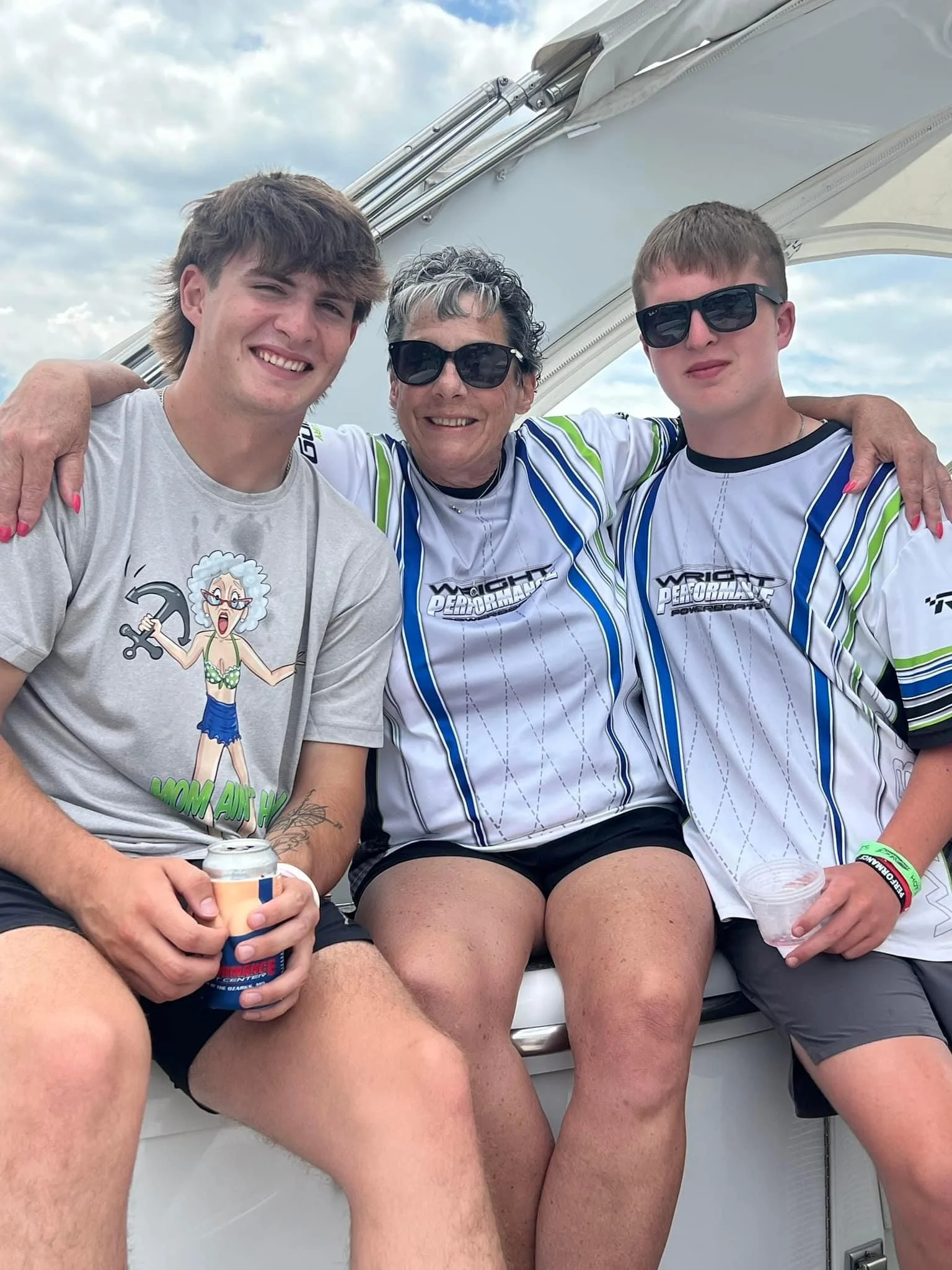 Three people sitting together outdoors on a boat, smiling, with two young men on either side of an older woman, all wearing sunglasses and casual clothing.