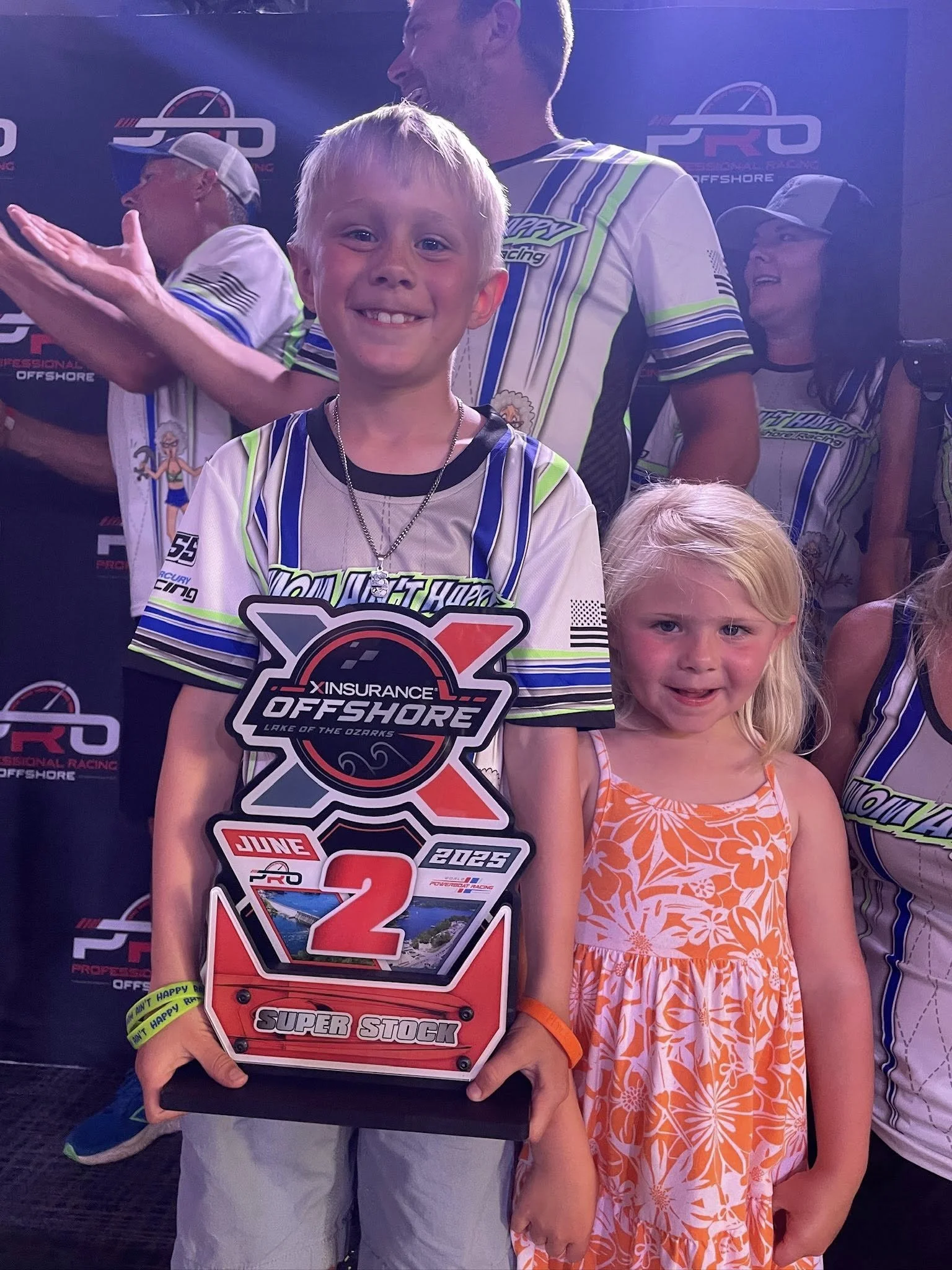 Smiling young boy holding a racing trophy at an off-road racing event with children and adults in racing shirts in the background.