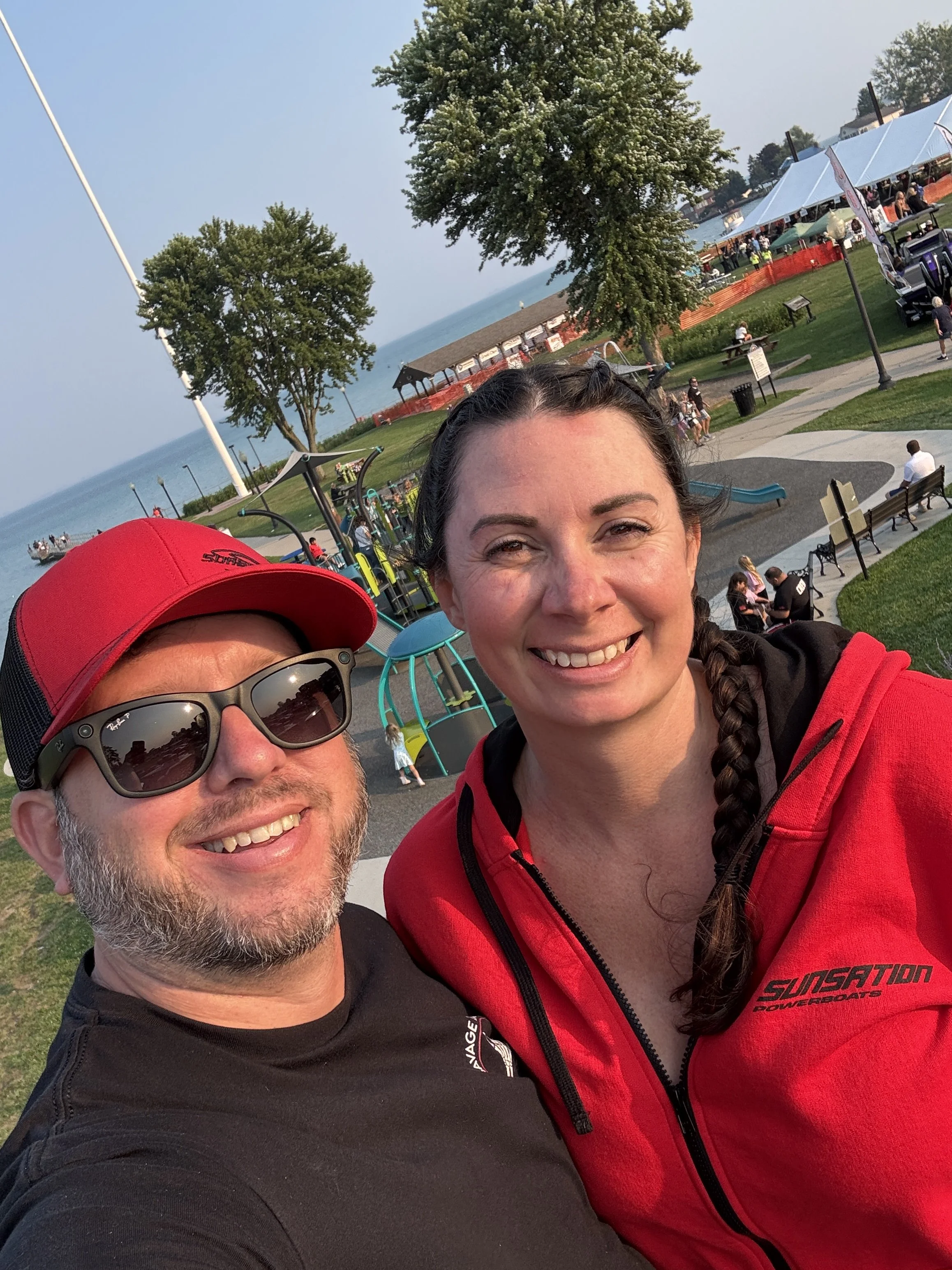 A smiling man and woman taking a selfie at a park near the water, with tents, trees, and other people in the background on a sunny day.
