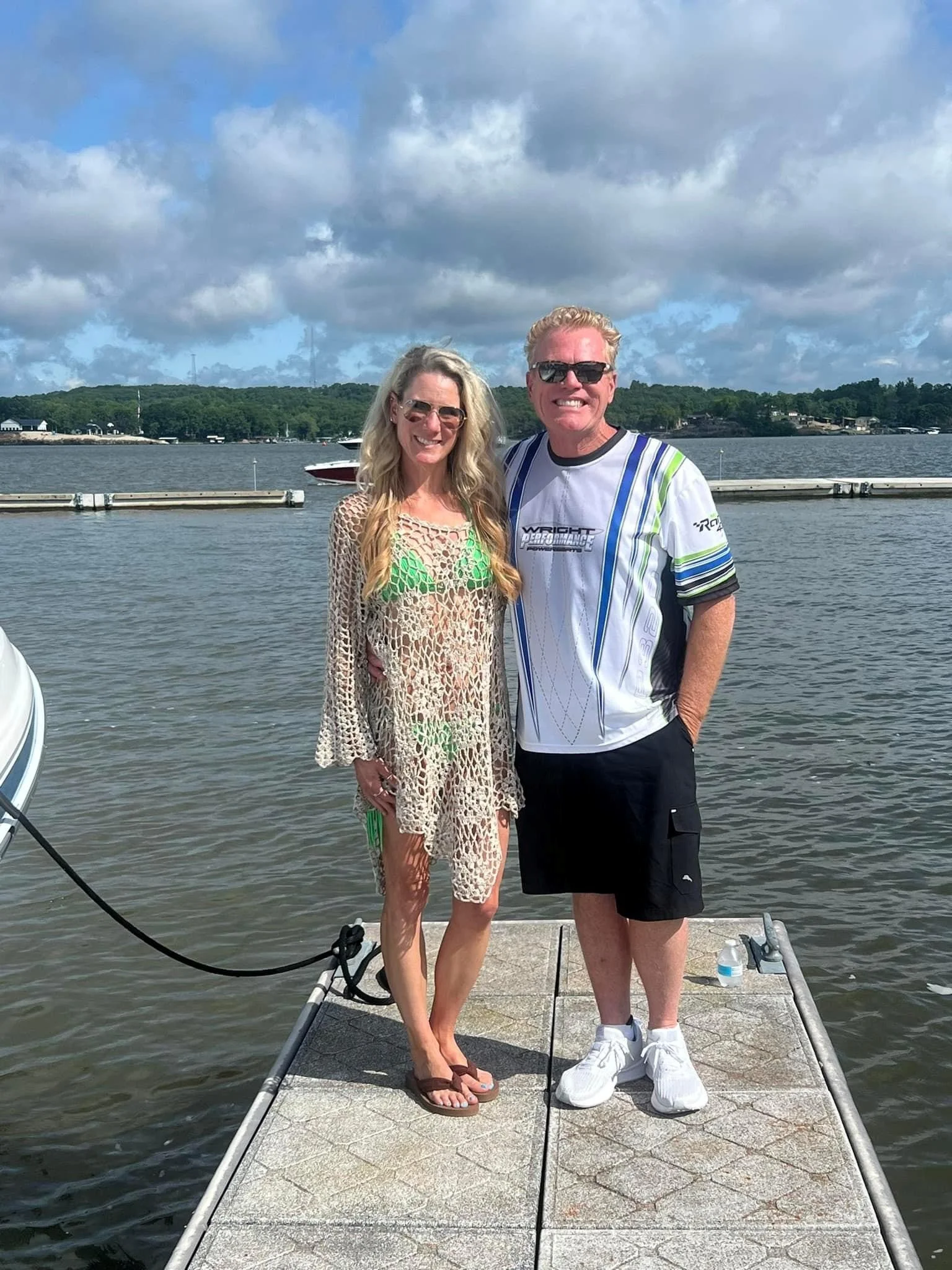 A man and woman standing on a dock by the water, smiling at the camera. The woman is wearing sunglasses, a crochet cover-up over a green swimsuit, and flip-flops. The man is wearing sunglasses, a white sports jersey, black shorts, and white sneakers.