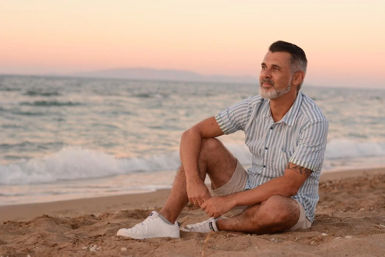 Hombre de cabello oscuro con barba, sentado en la playa al atardecer, usando camiseta de rayas y pantalones cortos.