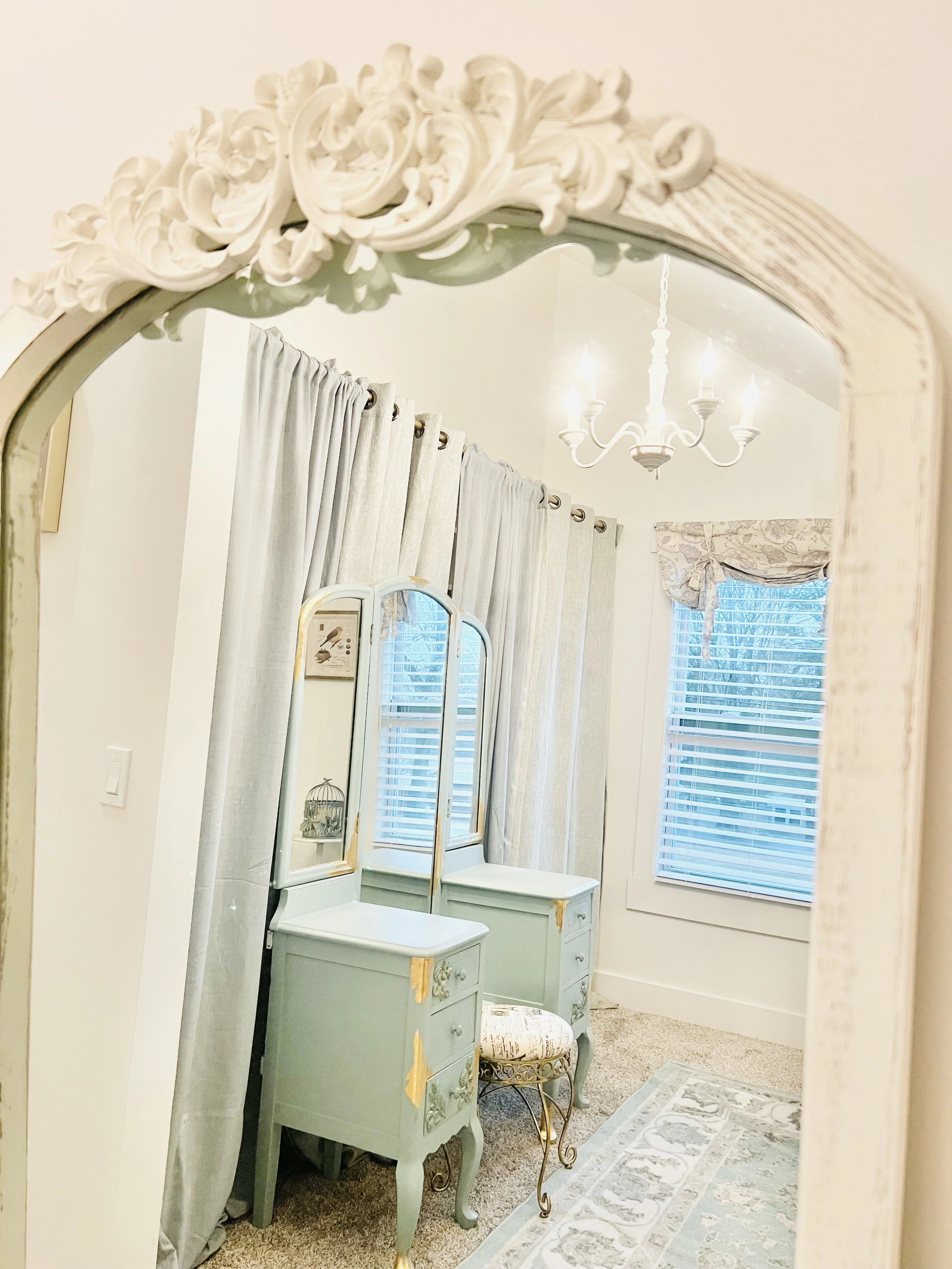 Decorative mirror reflecting a vanity with a stool, curtains, a window, and a chandelier in a bedroom.
