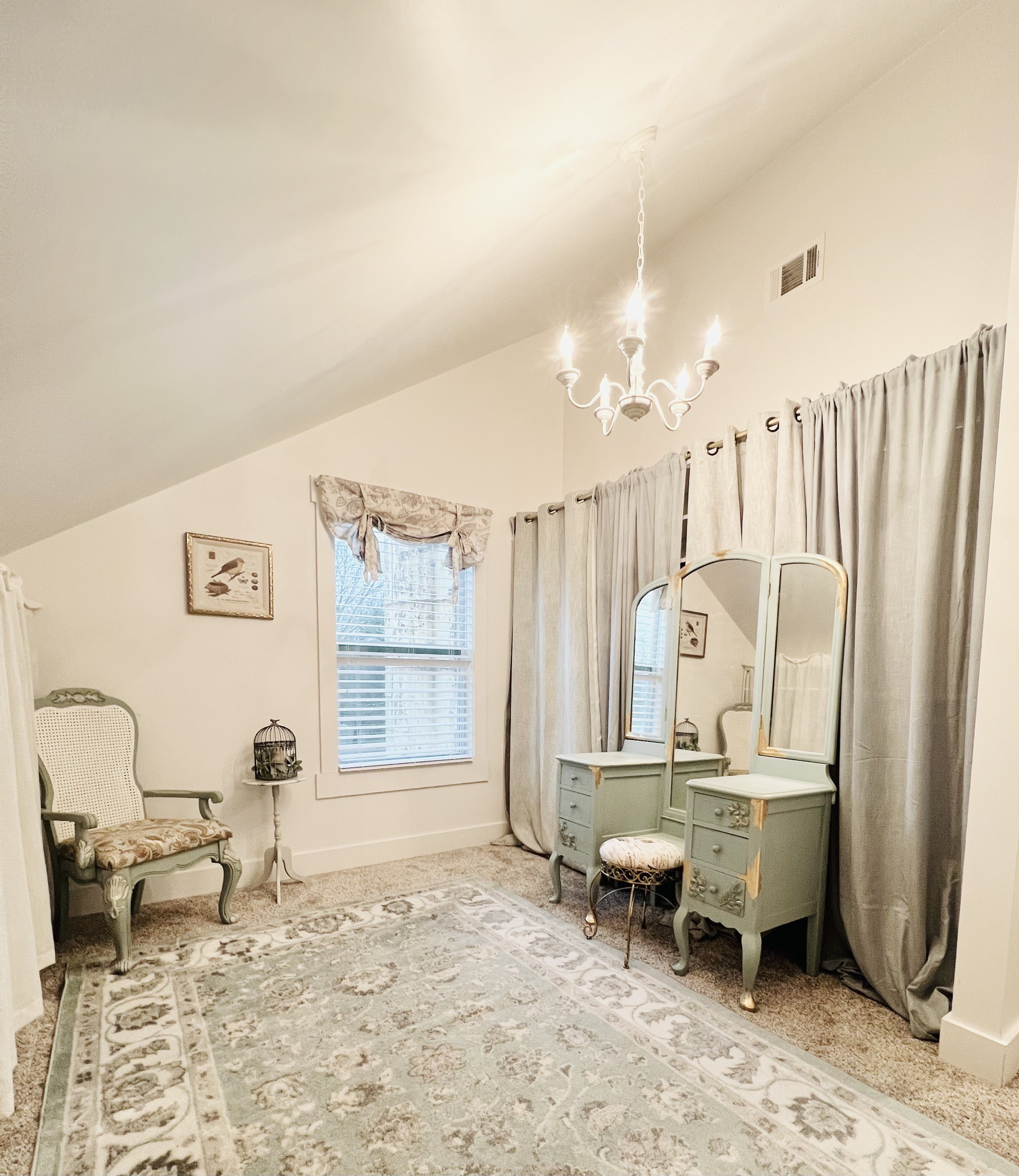 A vintage-style bedroom with a chandelier, a dresser with a large mirror, a small stool, a upholstered armchair, a side table with a birdcage, a window with valance, and beige curtains. The room has a patterned area rug and framed bird artwork.