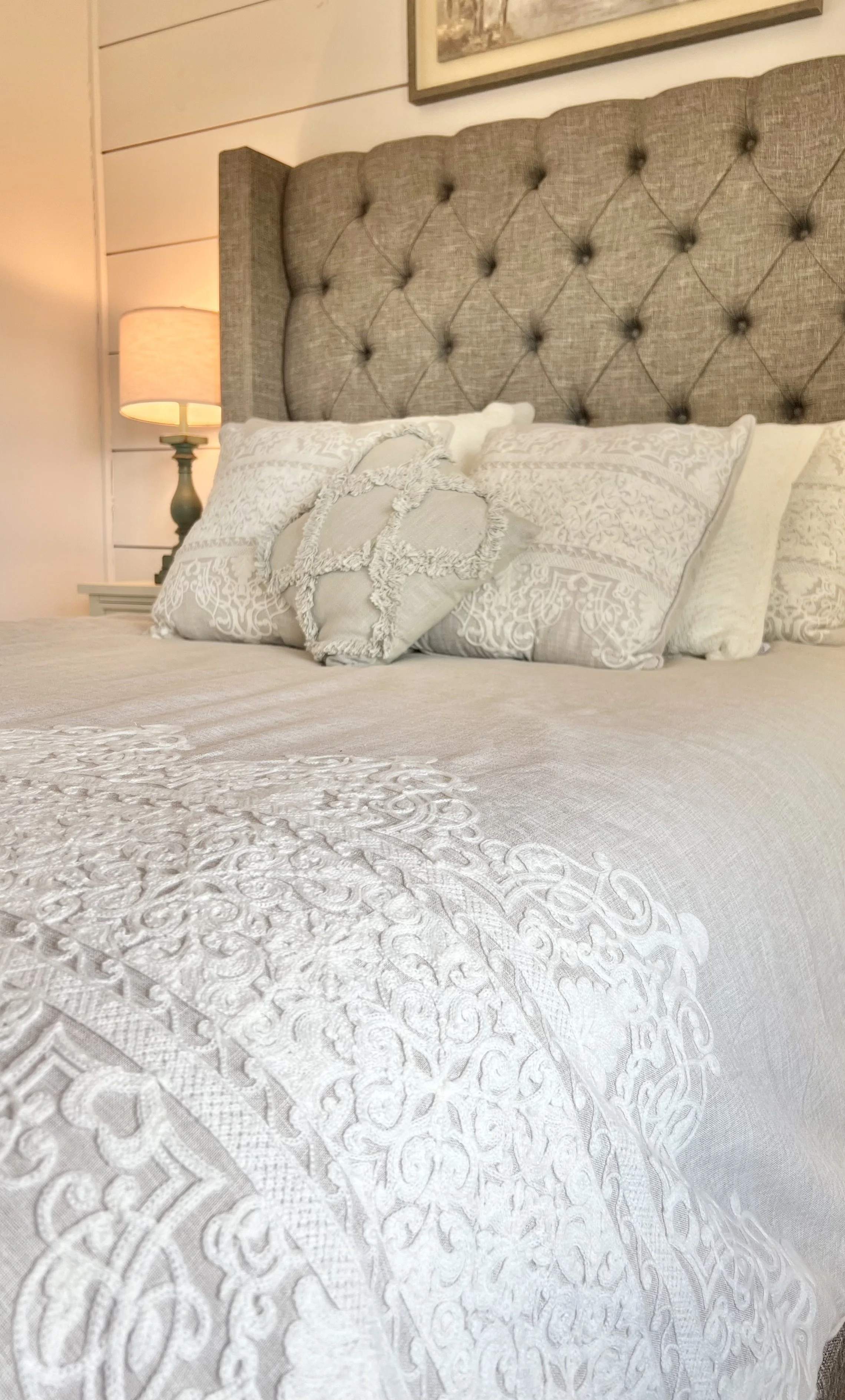 A bedroom with a bed that has a tufted beige headboard, white embroidered bedding, and decorative pillows, with a table lamp beside it.