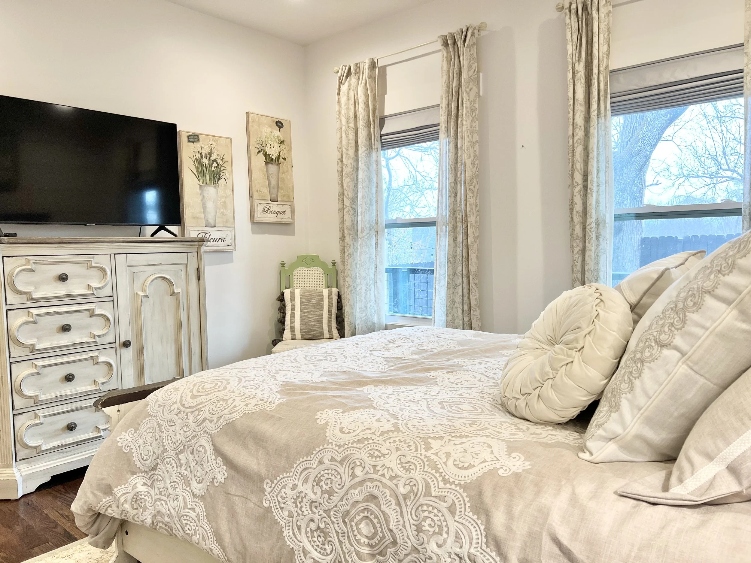 Cozy queen bedroom with layered neutral bedding, a vintage-style dresser, soft natural light, and French country charm at a Ball Ground, Georgia vacation rental.
