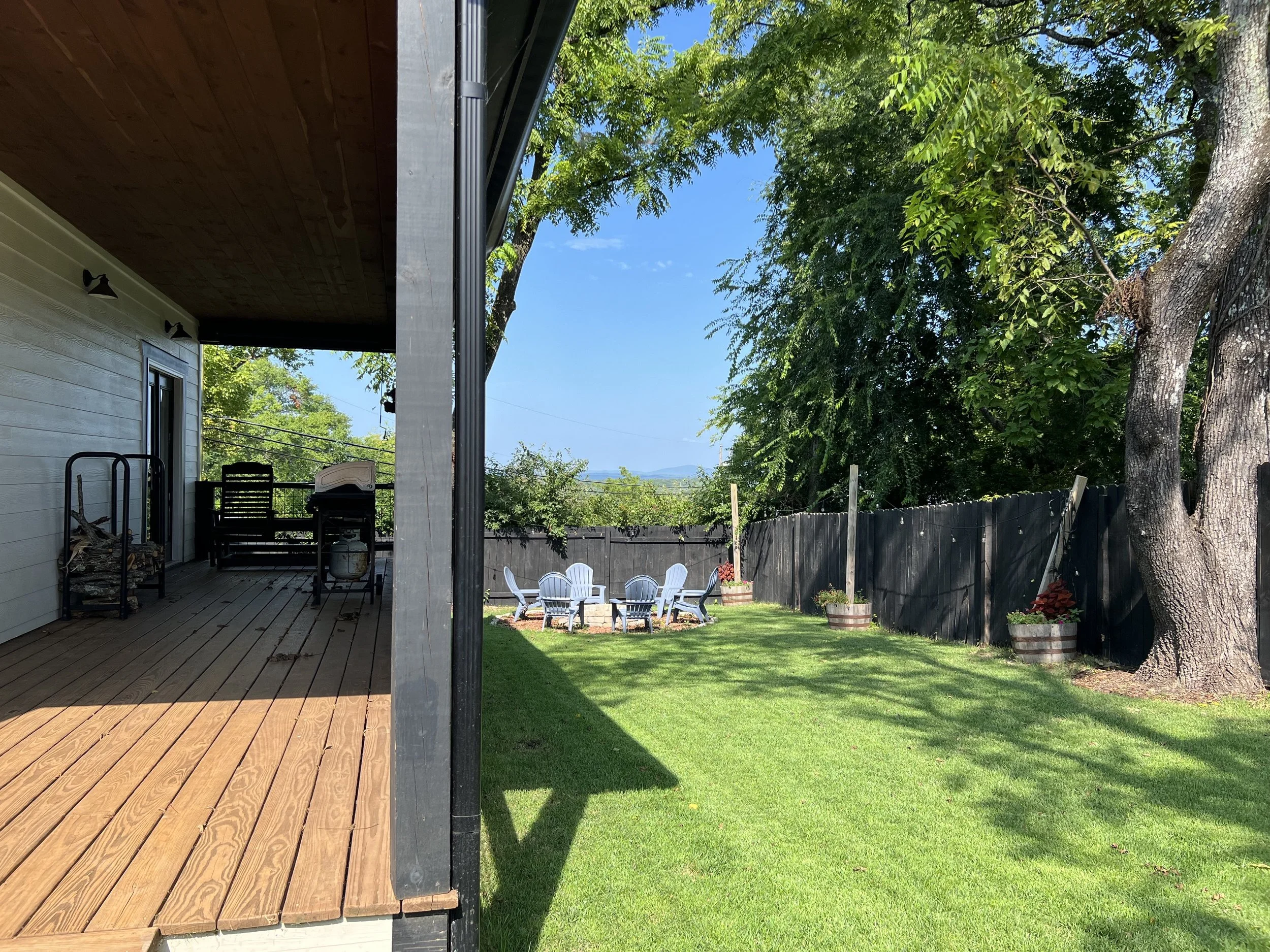 View of a backyard with a lawn, large trees, potted plants, a fenced perimeter, a stone fire pit with chairs around it, and a wooden deck attached to the house.