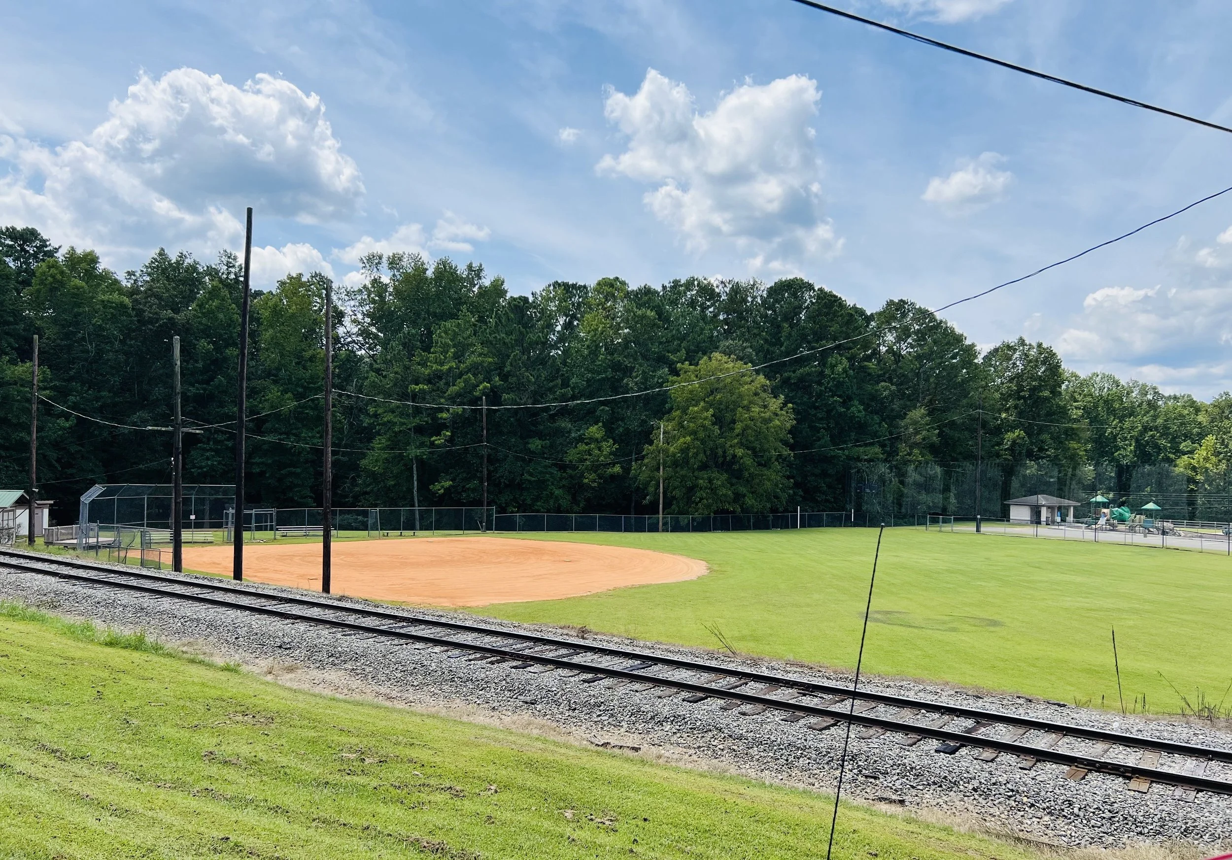 A baseball field near a railroad track with greenery and a blue sky with clouds.