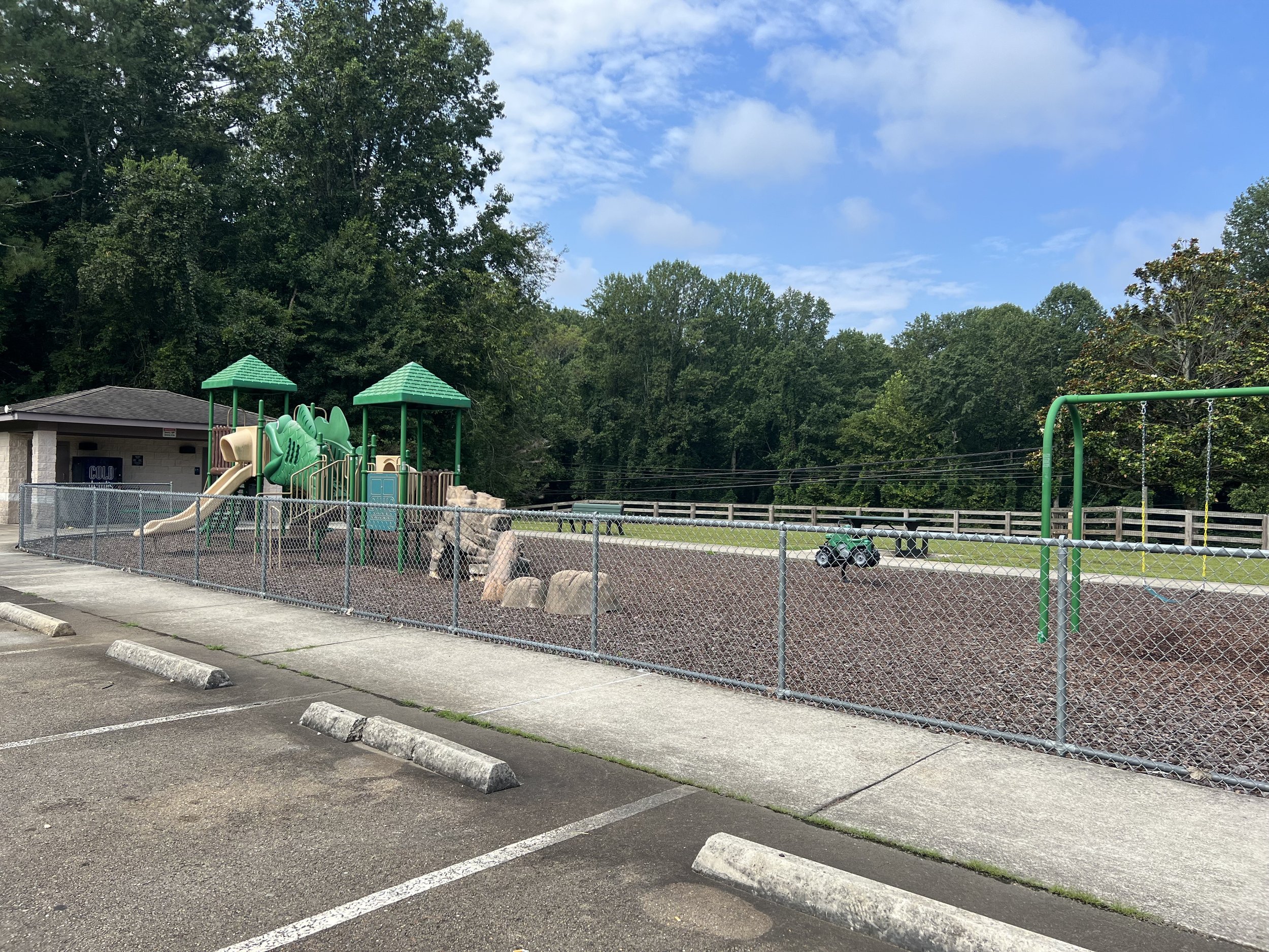 Empty playground and parking lot with a chain-link fence, surrounded by trees, under a partly cloudy sky.