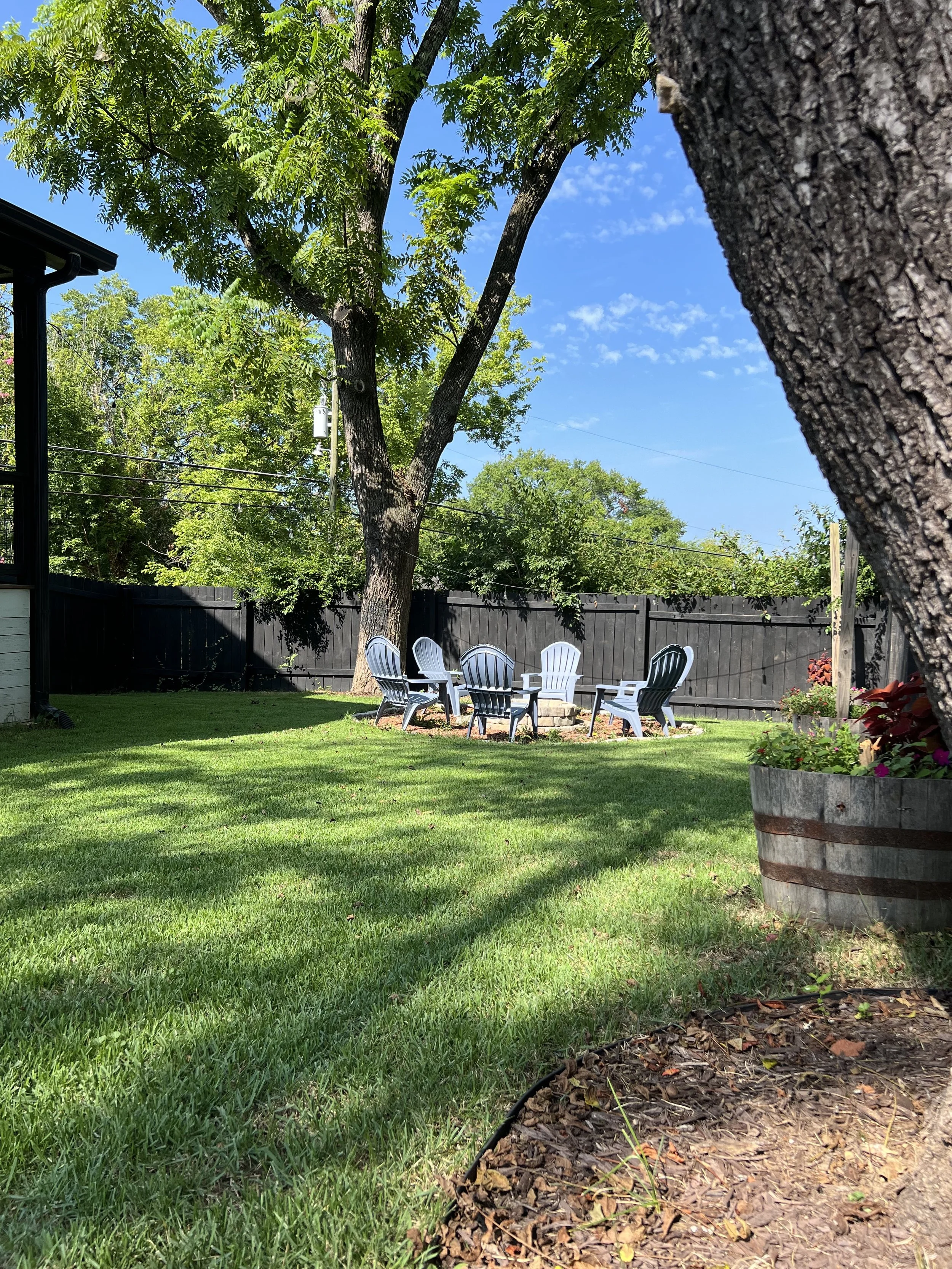 A backyard with a grassy lawn, large trees, a stone fire pit surrounded by white and green Adirondack chairs, a black wooden fence in the background, and a flower planter on the right.