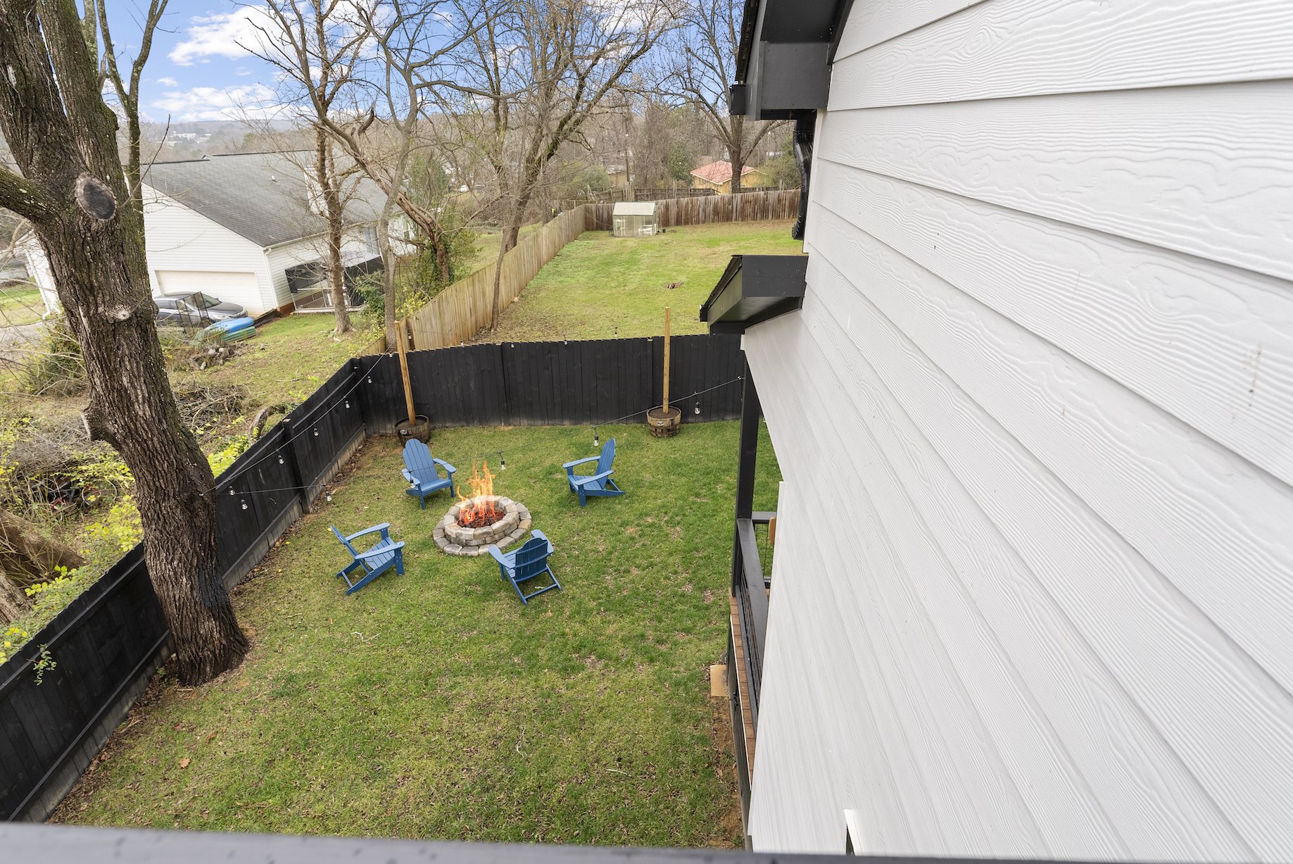 View from a house window showing a backyard with a firepit surrounded by five blue Adirondack chairs, a grassy lawn, a tall tree, wood fences, and neighboring houses.