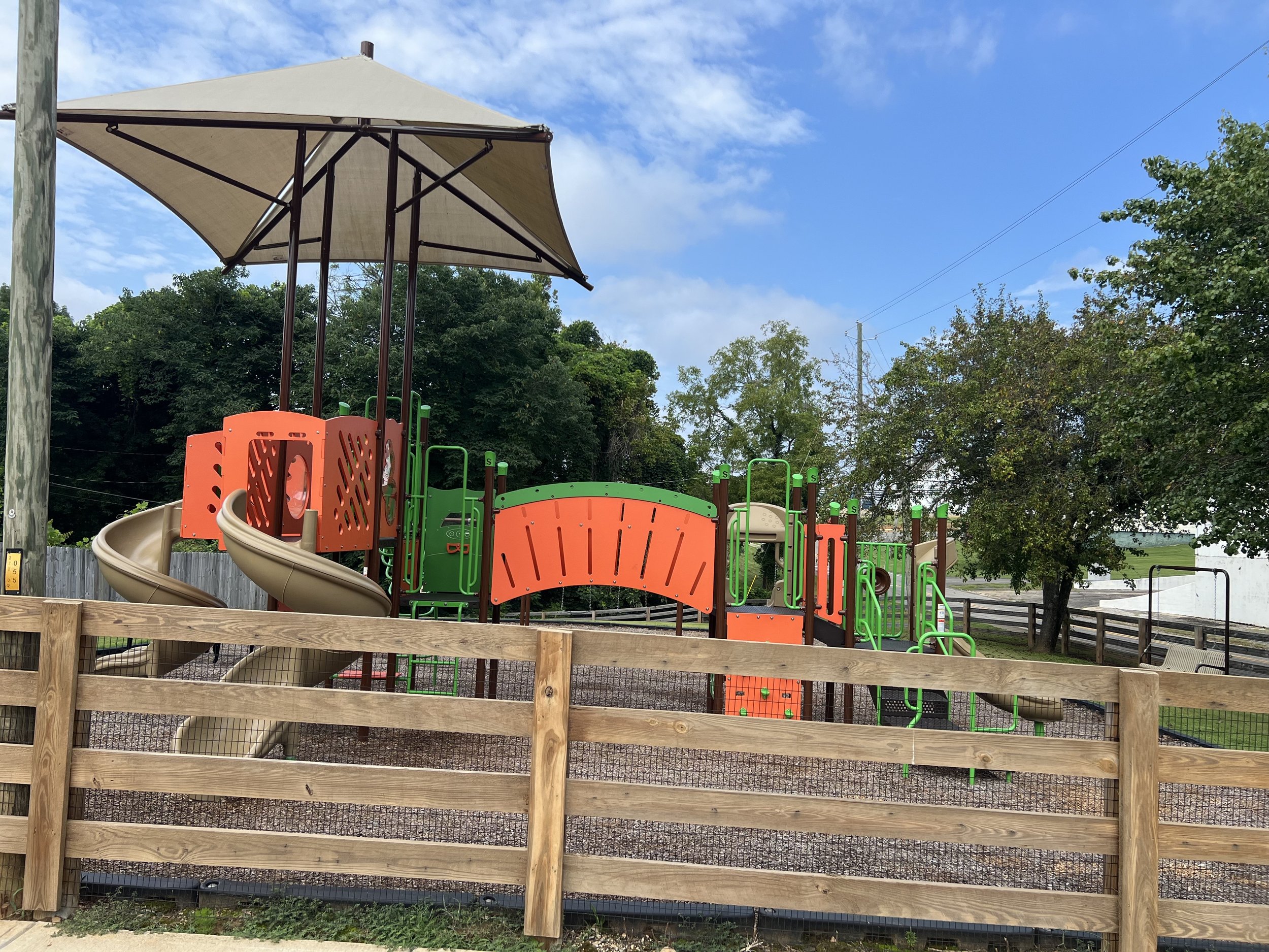 Colorful playground with slides, climbing structures, and a large beige umbrella, enclosed by a wooden fence, in a park with trees and a partly cloudy sky.