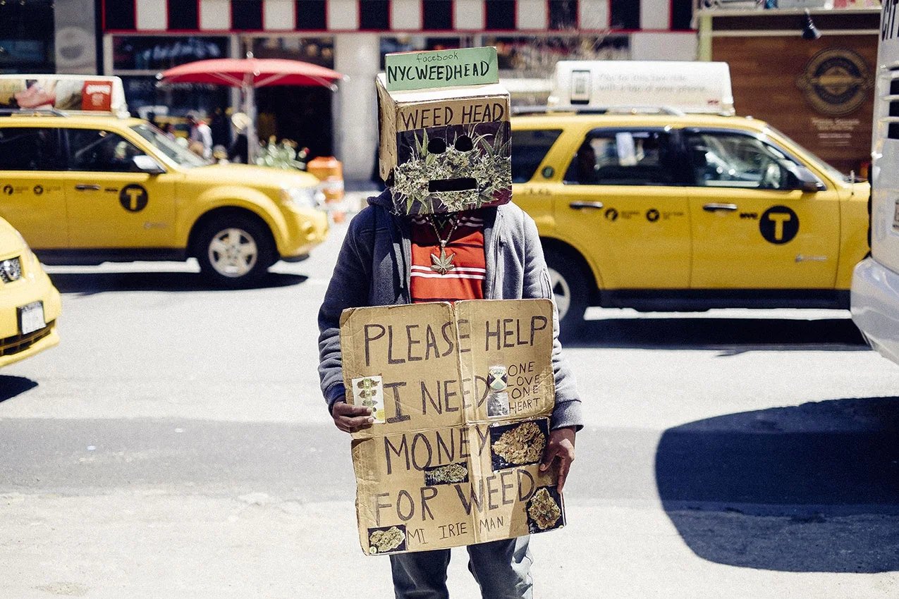 Person wearing a cardboard box with a face drawn on it, standing on a street with yellow taxis behind them, holding a large cardboard sign asking for money to buy weed.