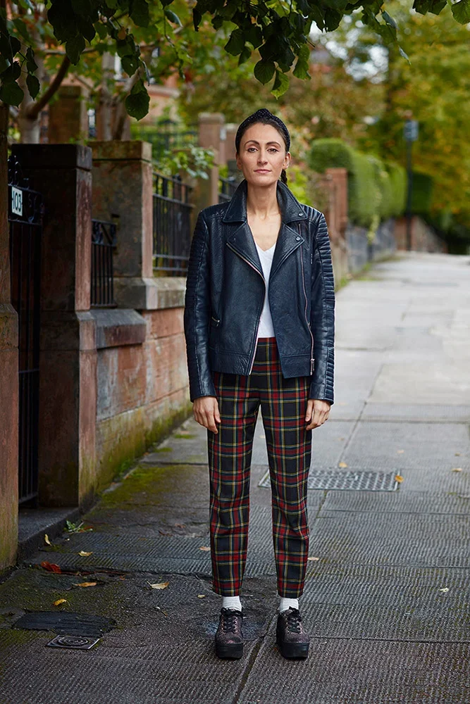 A woman standing on a sidewalk under a tree with green leaves, wearing a black leather jacket, white top, plaid pants, white socks, and black platform shoes.