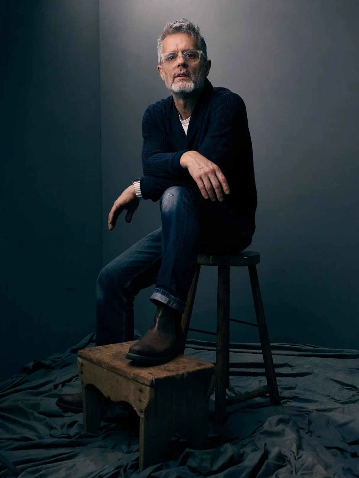 A man with glasses and gray hair sits on a wooden stool, wearing a dark sweater, jeans, and boots, against a plain dark background.