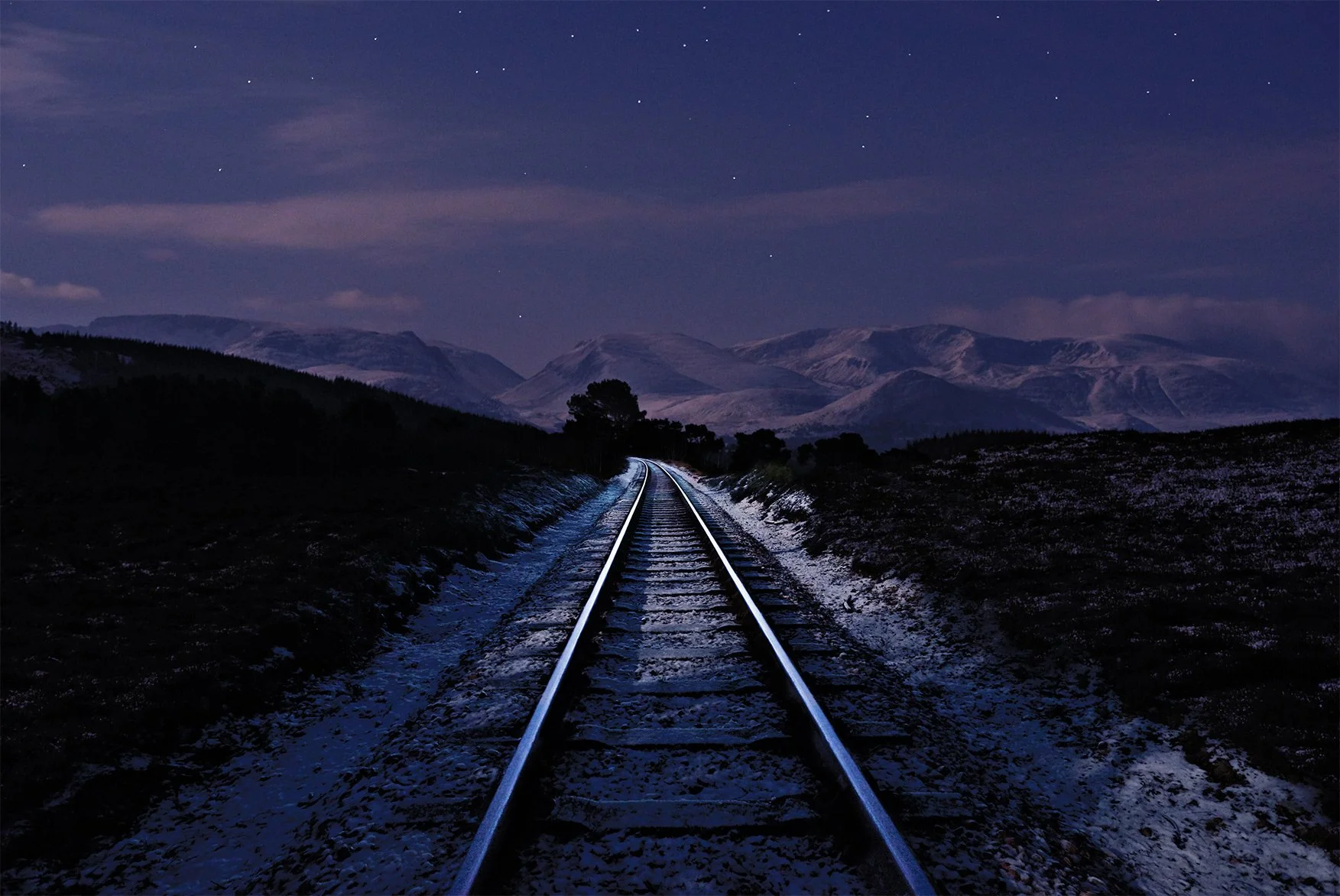 Night scene of railway tracks extending into the distance through a cold, snow-dusted landscape with mountains and a starry sky in the background.