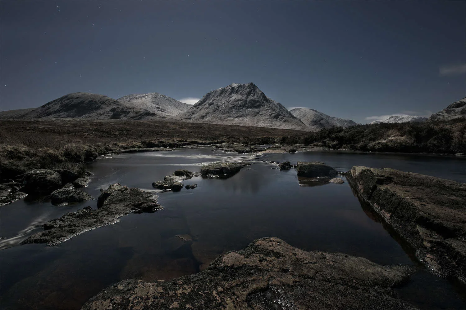 Nighttime landscape with a river flowing through rocks, mountain range with snow, and a starry sky