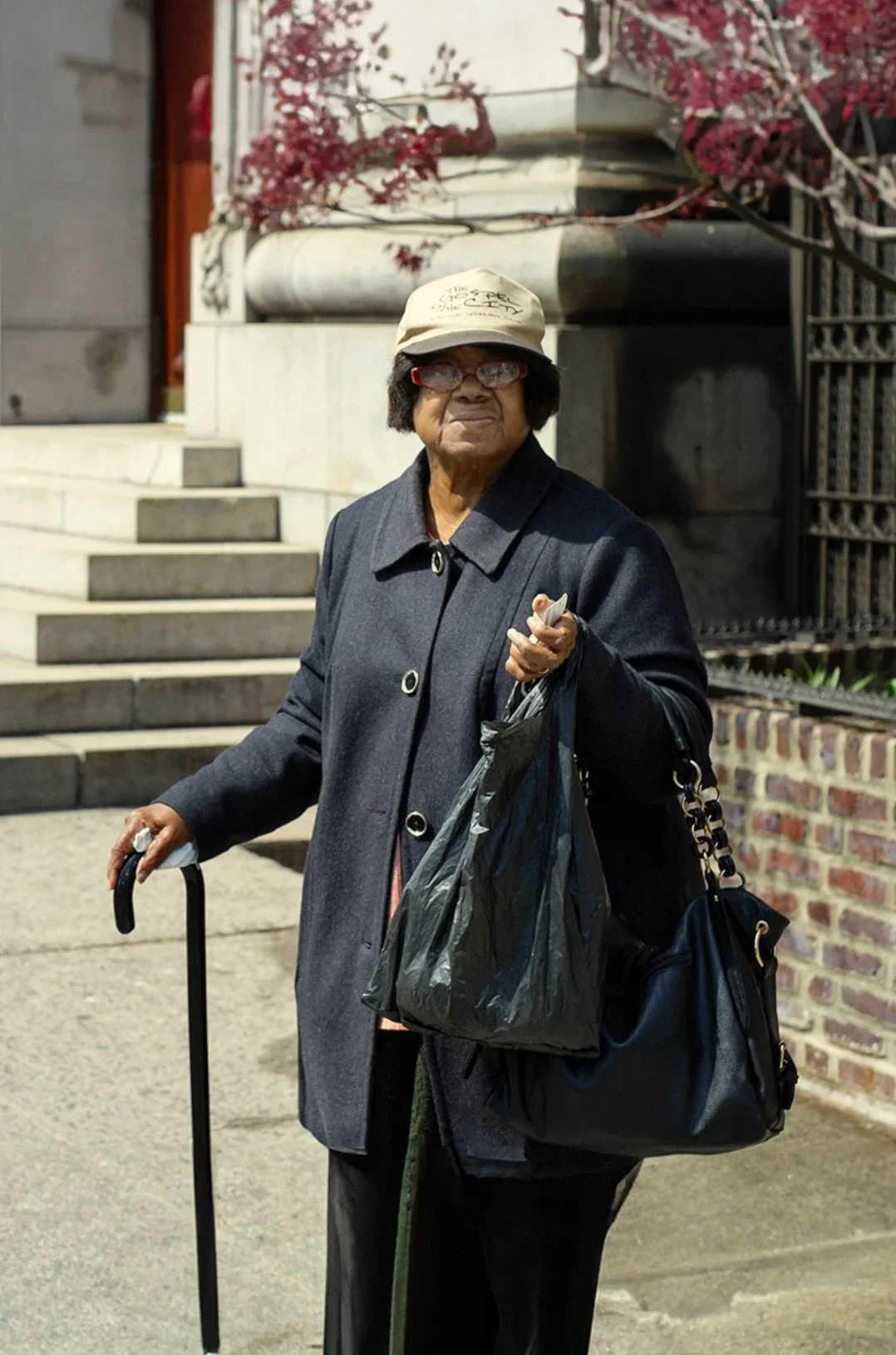 An elderly woman standing outside on a sidewalk, holding a cane in one hand and carrying black plastic bags and a black purse. She wears glasses, a beige hat, and a dark coat. In the background, there are stone steps, a brick wall, and some trees wit
