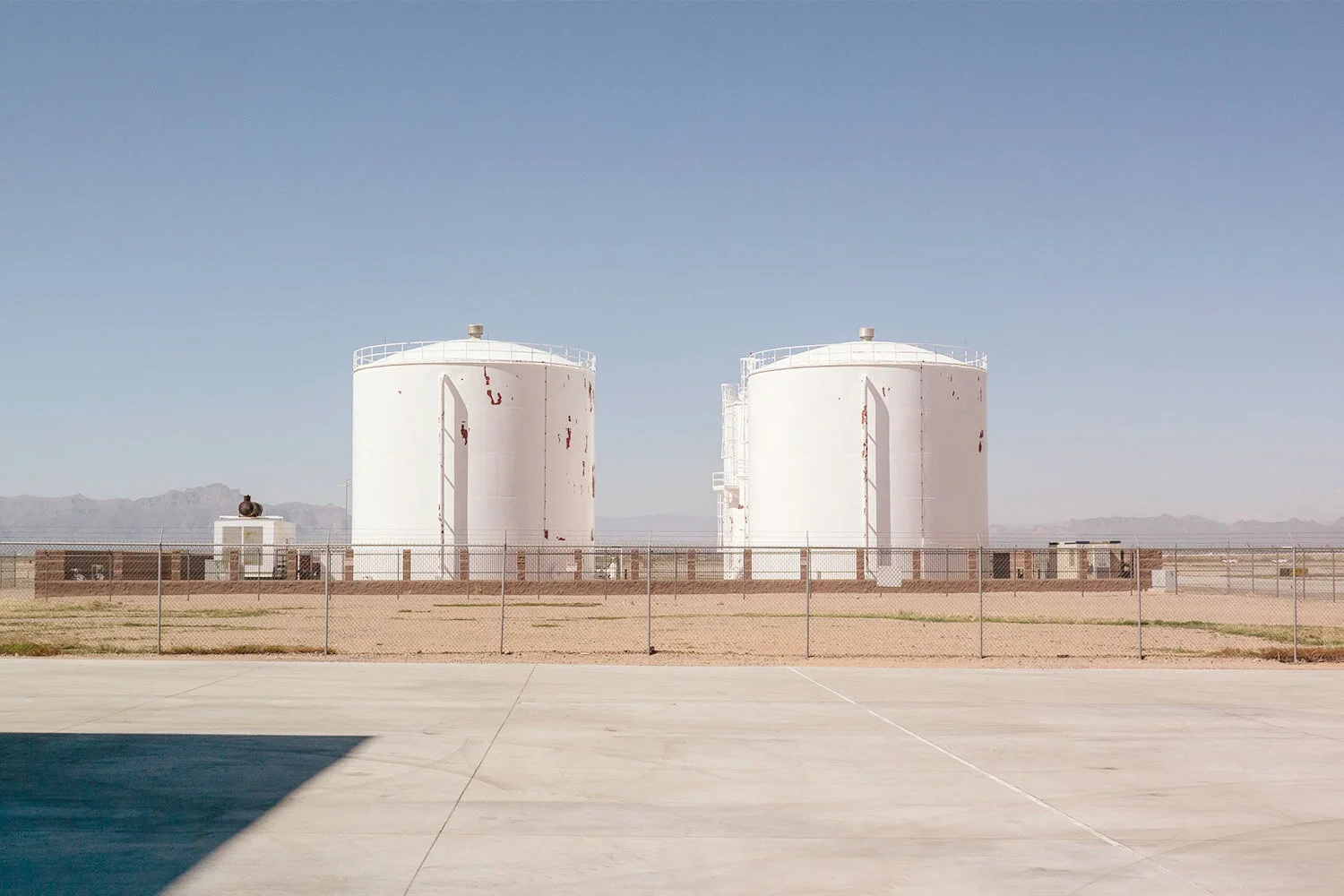 Two large white industrial storage tanks in a desert area, surrounded by a chain-link fence, with clear blue sky and distant mountains in the background.
