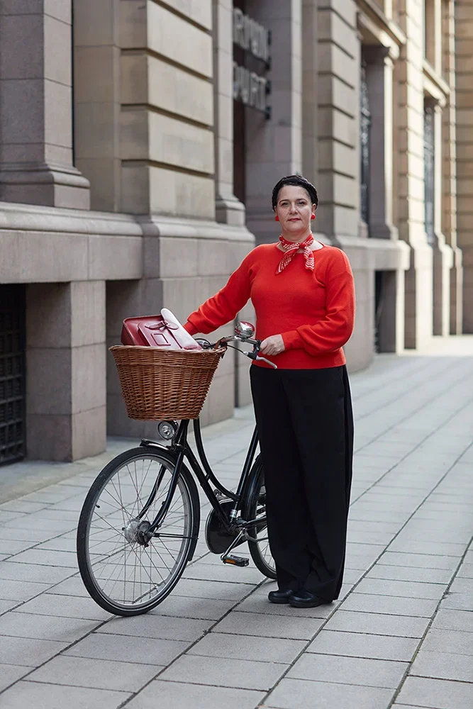 Woman standing on city sidewalk holding a bicycle with a basket containing a red shopping bag and a white item, in front of a stone building with large windows.