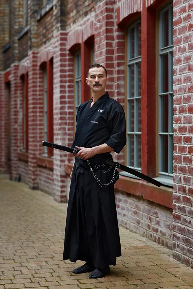 Man dressed in black martial arts uniform with sword, standing on brick sidewalk outside red brick building with windows.