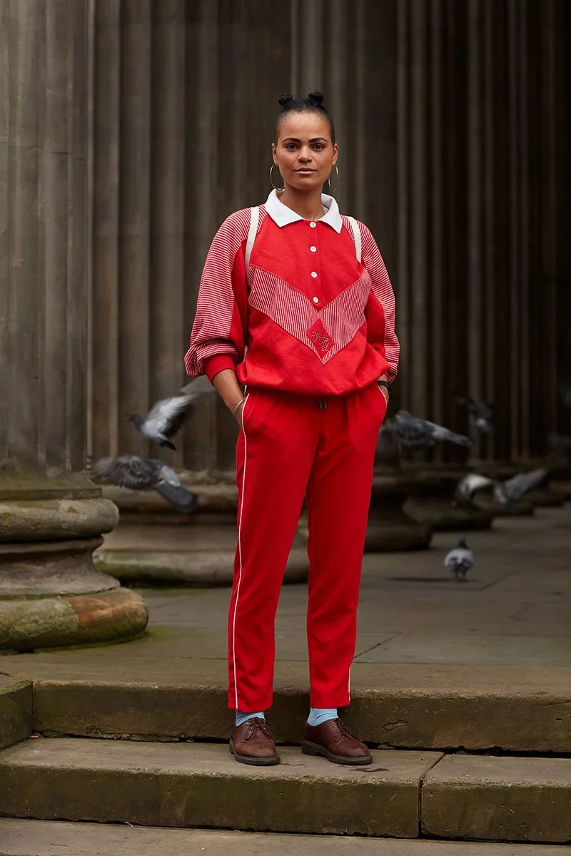 Young woman standing outdoors in front of columns, wearing a red and white athletic tracksuit, with pigeons nearby.