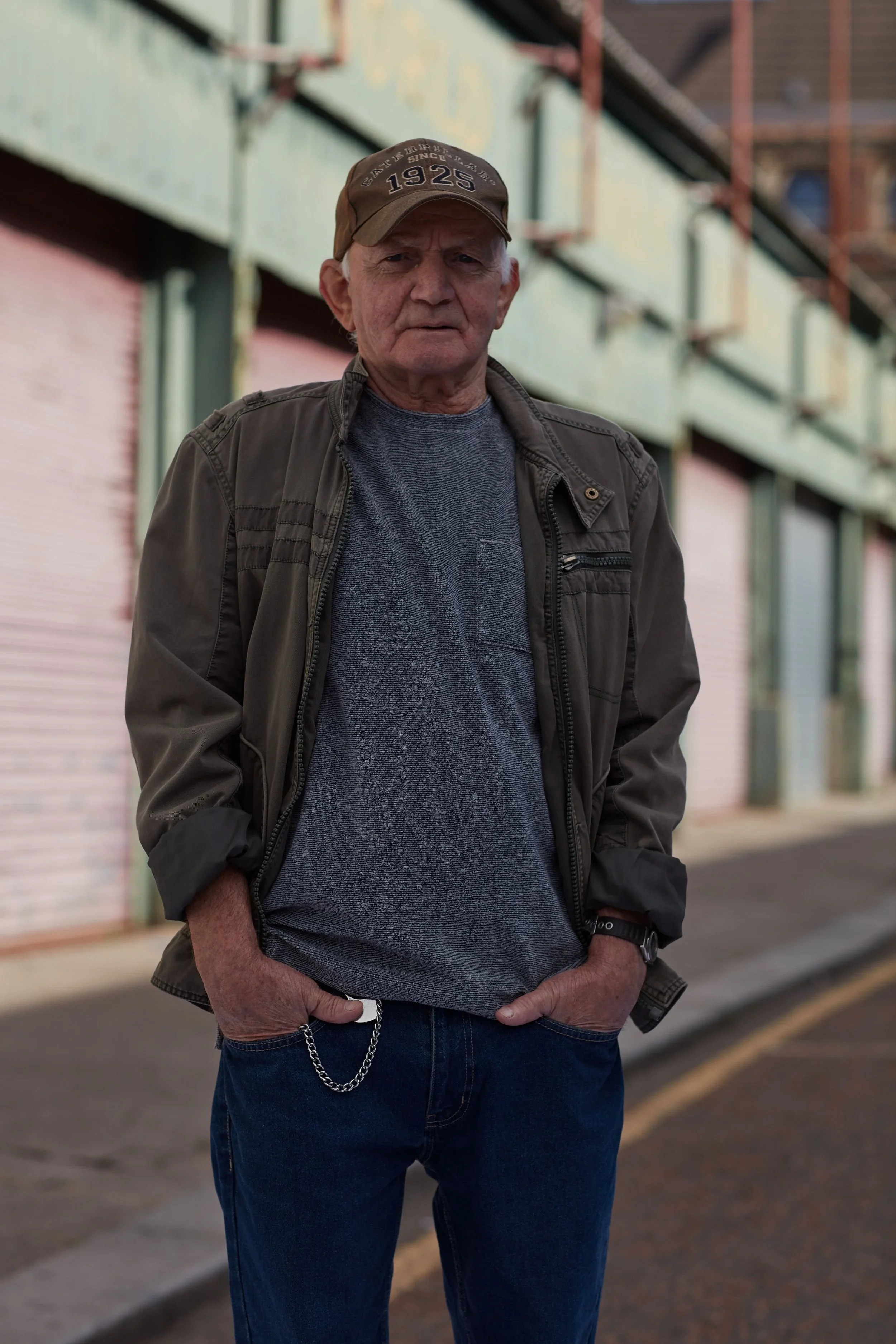An elderly man with gray hair, wearing a cap, gray t-shirt, and brown jacket, stands with his hands in his pockets on a city street with closed buildings in the background.