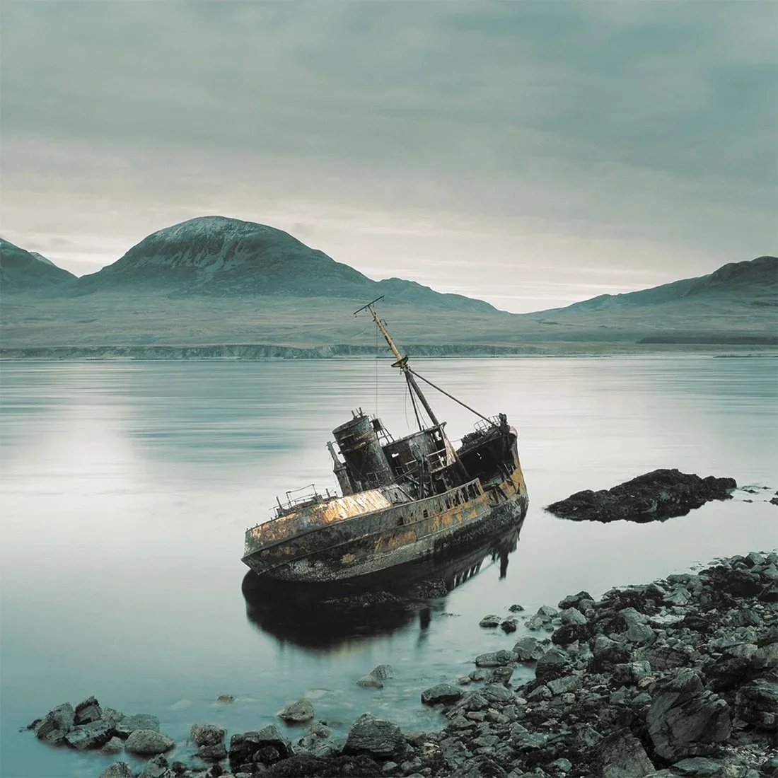 A rusted, abandoned shipwreck leaning to one side in calm water near a rocky shoreline, with mountains and an overcast sky in the background.