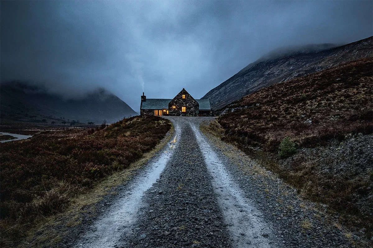 A stone house with lit windows at the end of a gravel driveway, surrounded by hills and mountains under a cloudy, dark sky.