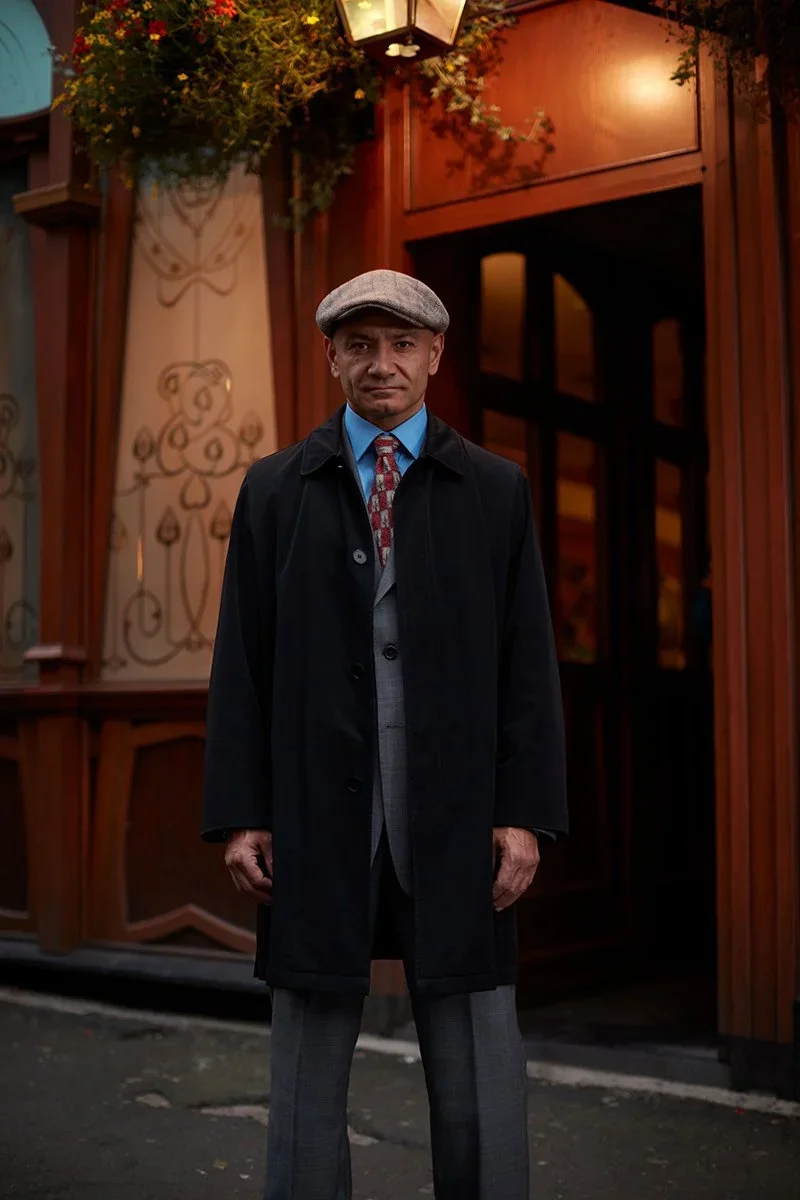A man standing outside in front of a wooden building with decorative ironwork and greenery, dressed in a gray cap, dark coat, suit, and tie.