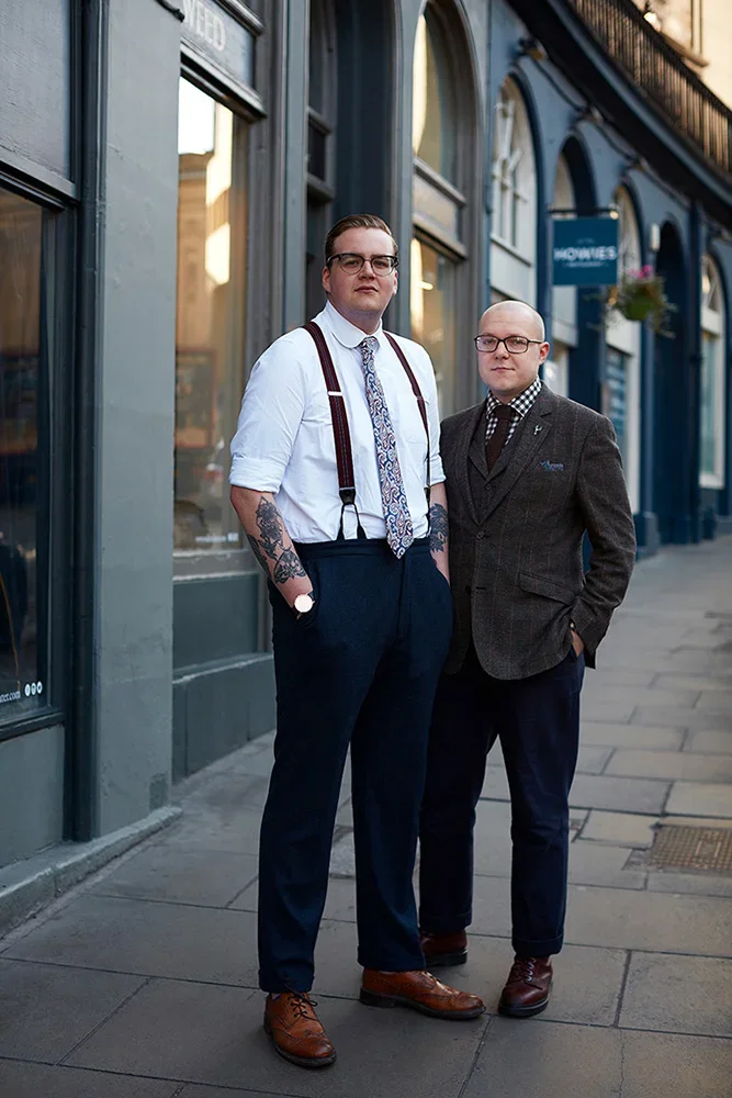 Two men standing on a city sidewalk in front of a building with large windows and a blue exterior. The man on the left has tattoos, glasses, and is dressed in a white shirt, patterned tie, suspenders, and dark trousers. The man on the right is bald, 