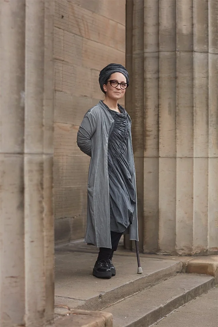 Woman with short gray hair and glasses standing on a city sidewalk next to classical stone columns, dressed in a gray coat and dress, with a cane.