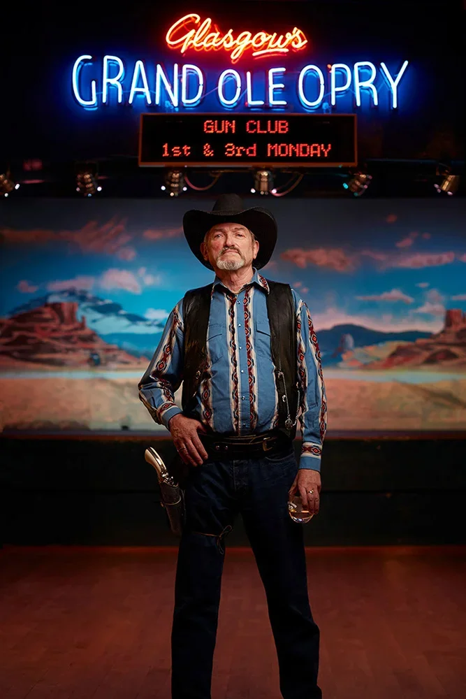 A man dressed as a cowboy with a black hat, striped shirt, and black vest stands in front of a desert landscape backdrop at Glascow's Grand Ole Opry. Neon signs above show "Glascow's GRAND OLE OPRY" and a digital sign advertises a gun club meet on th