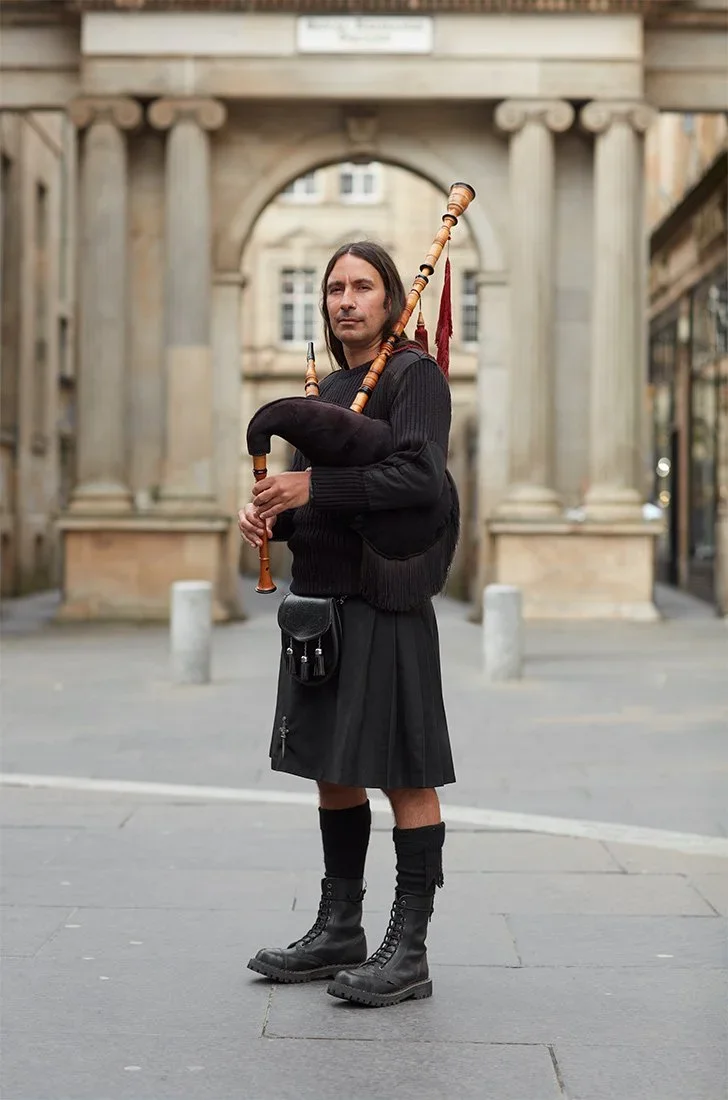 A man dressed in traditional Scottish attire, including a kilt, black boots, and a black sweater, stands on a city street holding a bagpipe. The background features an ornate stone archway.