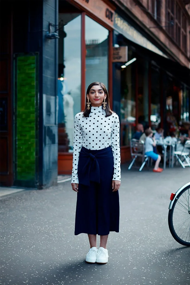 A young woman standing on a city sidewalk in front of a restaurant or café with outdoor seating. She is wearing a white polka dot long-sleeve top, navy wide-leg pants with a tied waist, and white sneakers. She has dark hair, gold earrings, and is pos
