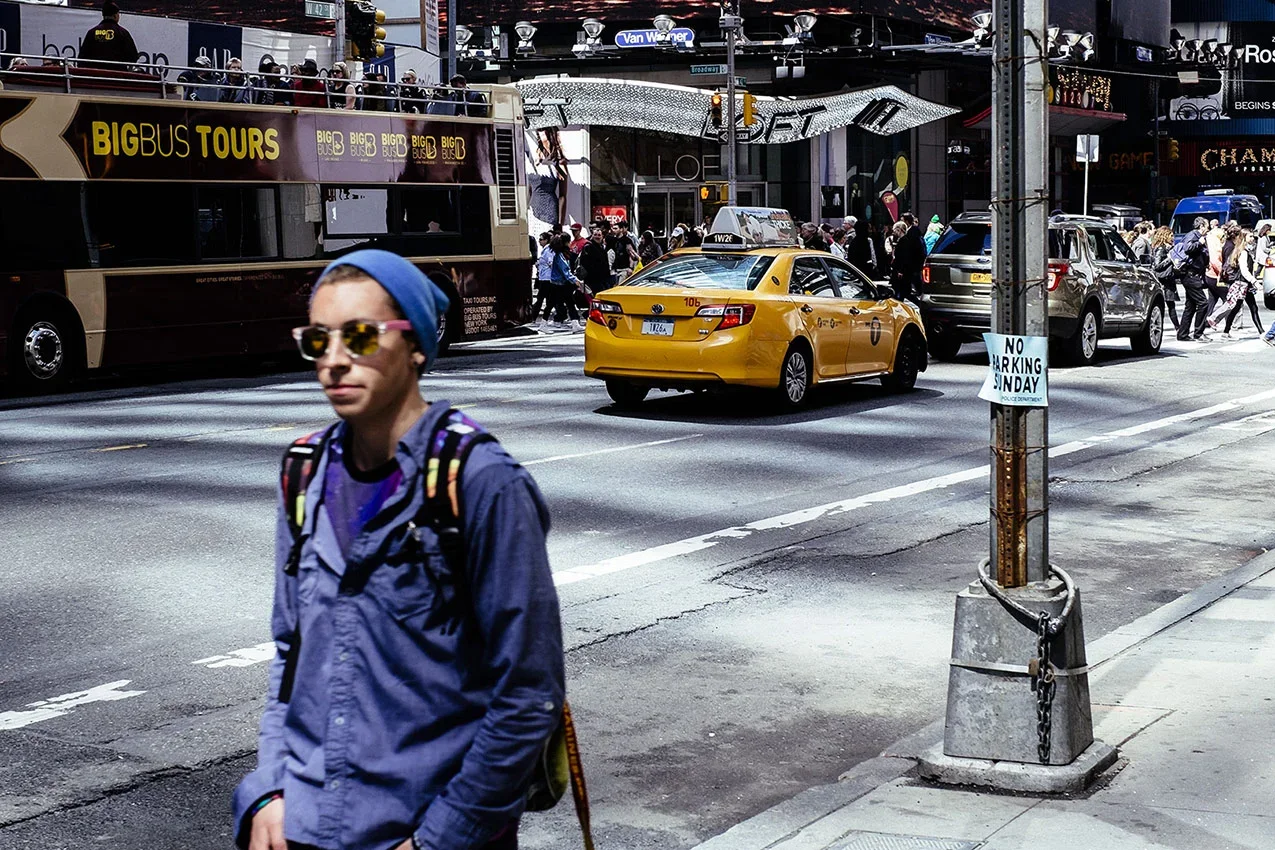 A busy city street scene with a man in the foreground wearing sunglasses, a blue beanie, a backpack, and a jacket. In the background, there are yellow taxis, a tour bus, and many pedestrians crossing the street. A sign on a lamppost reads 'No Parking