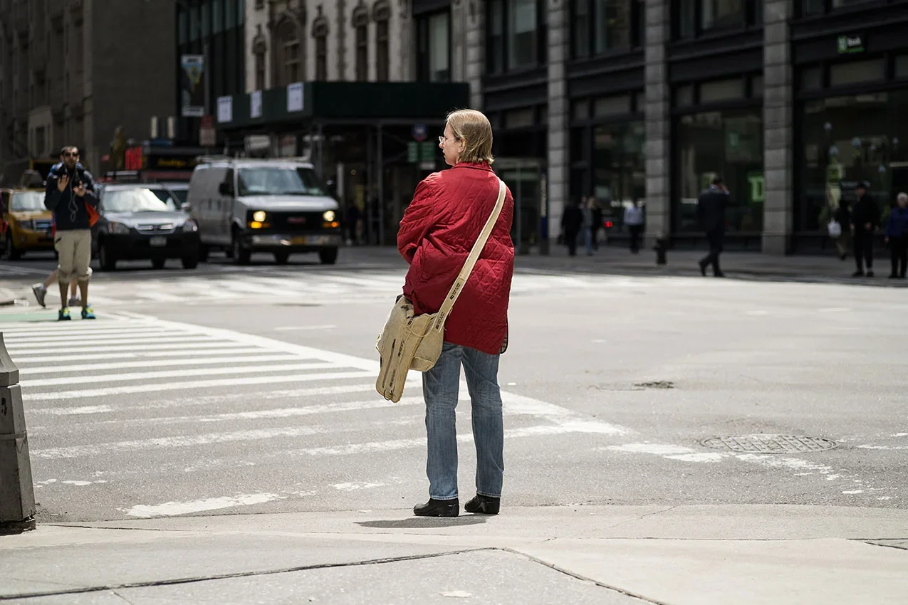 A woman in a red quilted jacket and jeans waits at a crosswalk in a city street, carrying a tan bag with a long strap.
