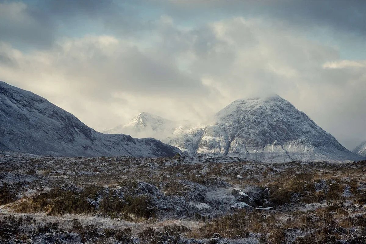 Snow-capped mountains with cloudy sky and rugged terrain in the foreground.