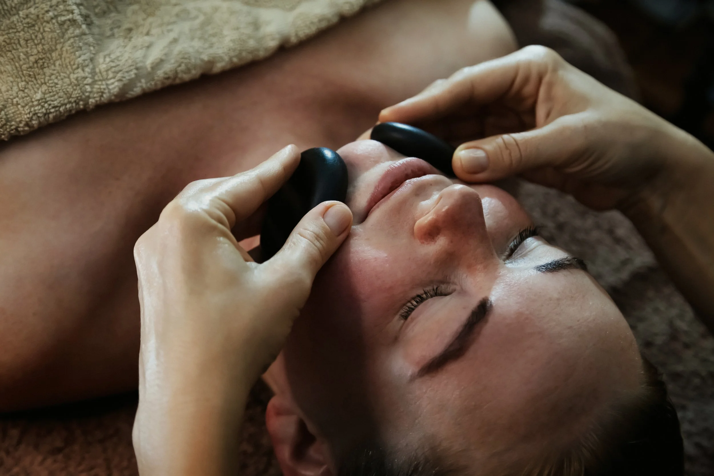 A person receiving a hot stone massage treatment, lying on a massage table with stones placed on their face, covered with a towel.