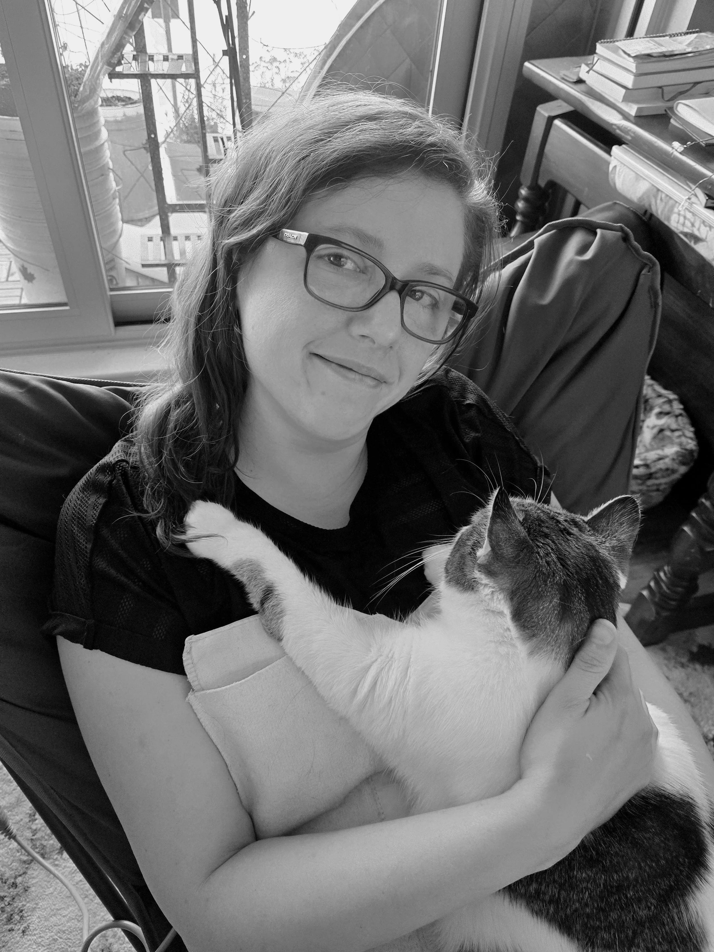 A woman with glasses sitting on a chair, holding and cuddling a cat with black and white fur, near a window with a view of an outdoor balcony.