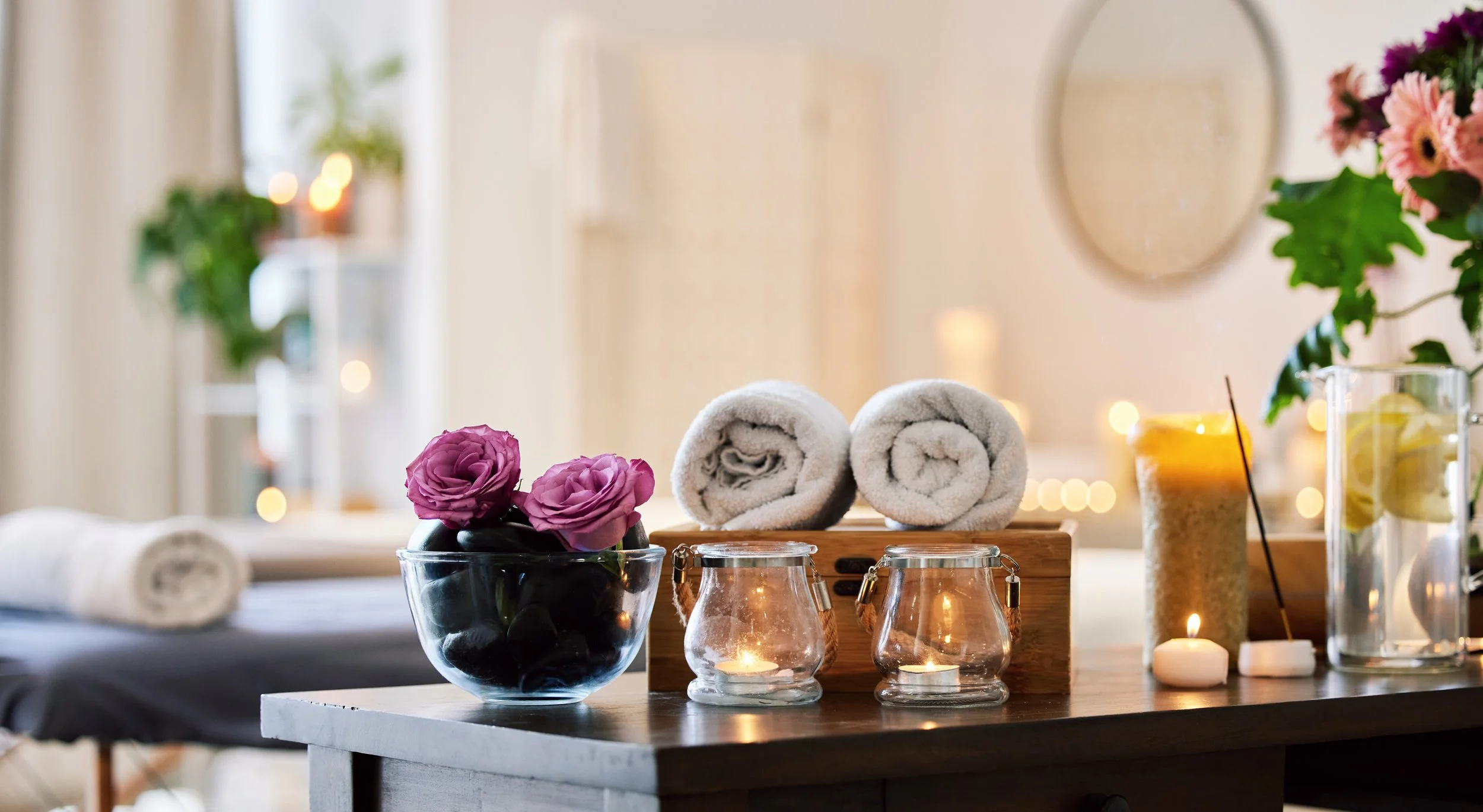 Spa or massage room with rolled white towels, pink roses in a black bowl, candles, and flower arrangements on a dark wooden table.